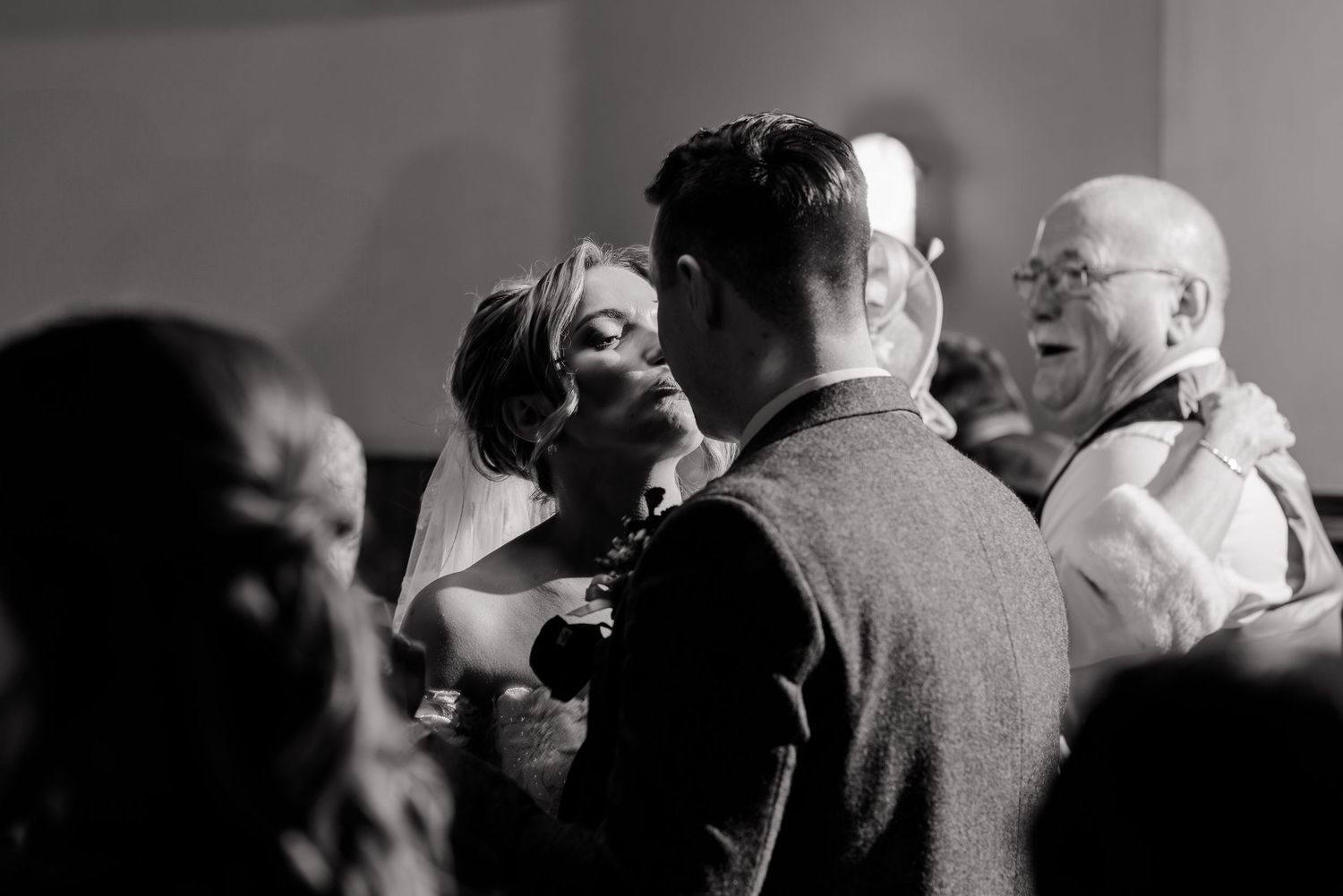 Black and white candid photo of bride and groom dancing with guests at wedding reception.