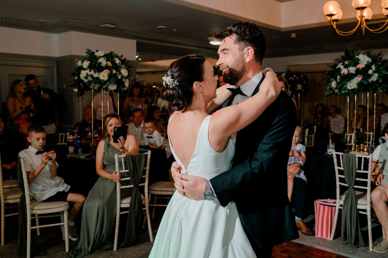 Bride in a white gown and groom in a dark suit share a close first dance while wedding guests watch from tables with floral centerpieces.