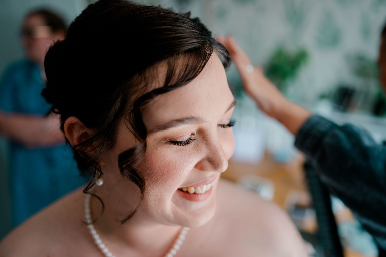 Bride smiling joyfully while having her hair styled on her wedding day, wearing pearl necklace and earrings.