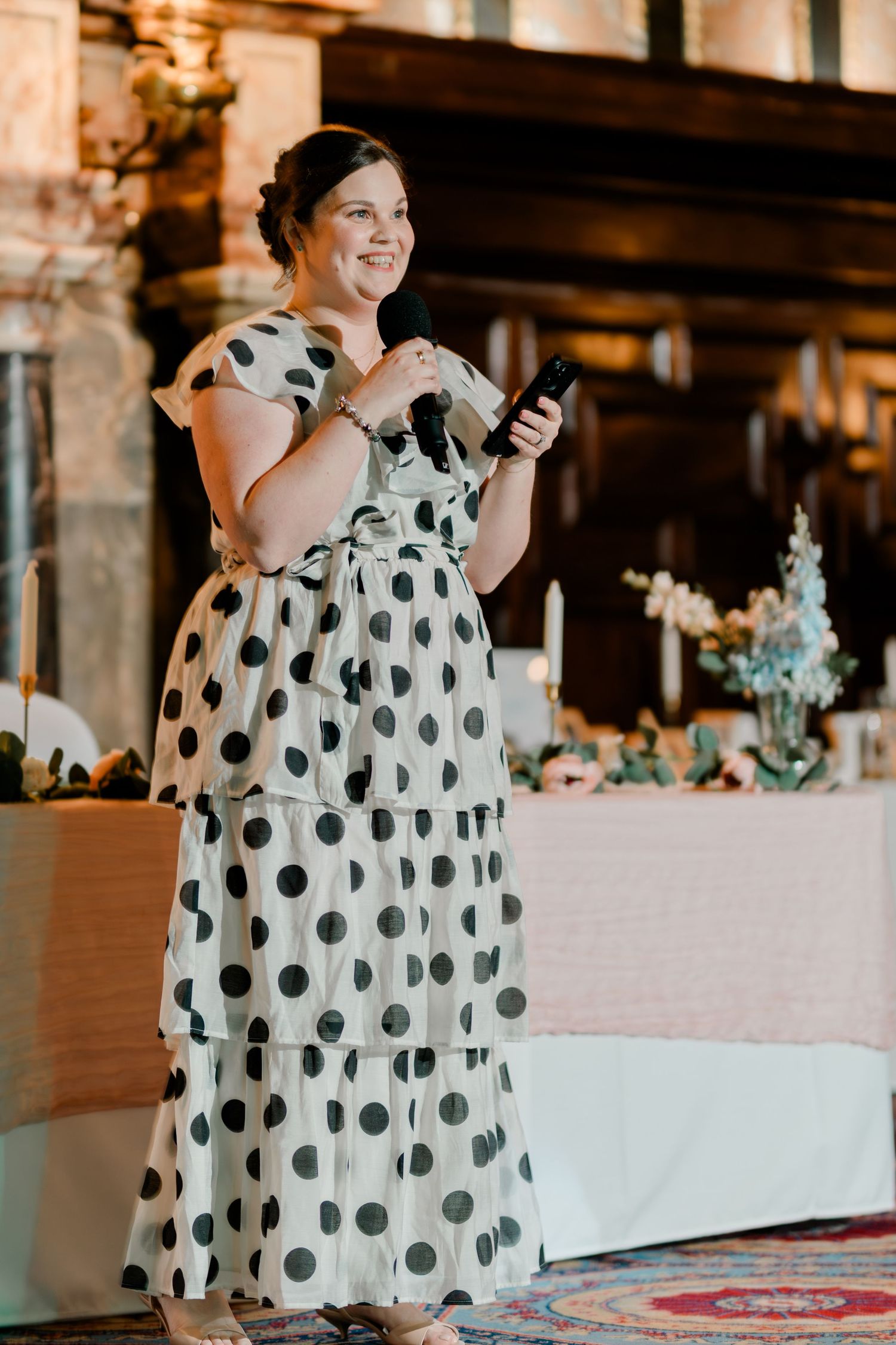 Smiling woman in a white and black polka dot tiered dress giving a speech at a wedding reception.
