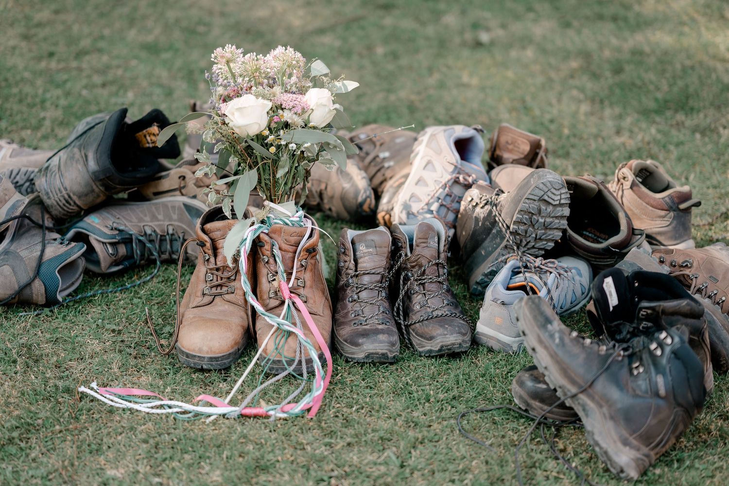 Multiple worn hiking boots arranged on grass surrounding a bouquet of white roses in an outdoor rustic wedding setting.