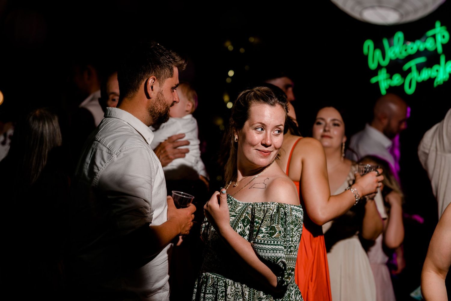 Guests dancing and socializing at a wedding reception with a neon jungle sign in the background.