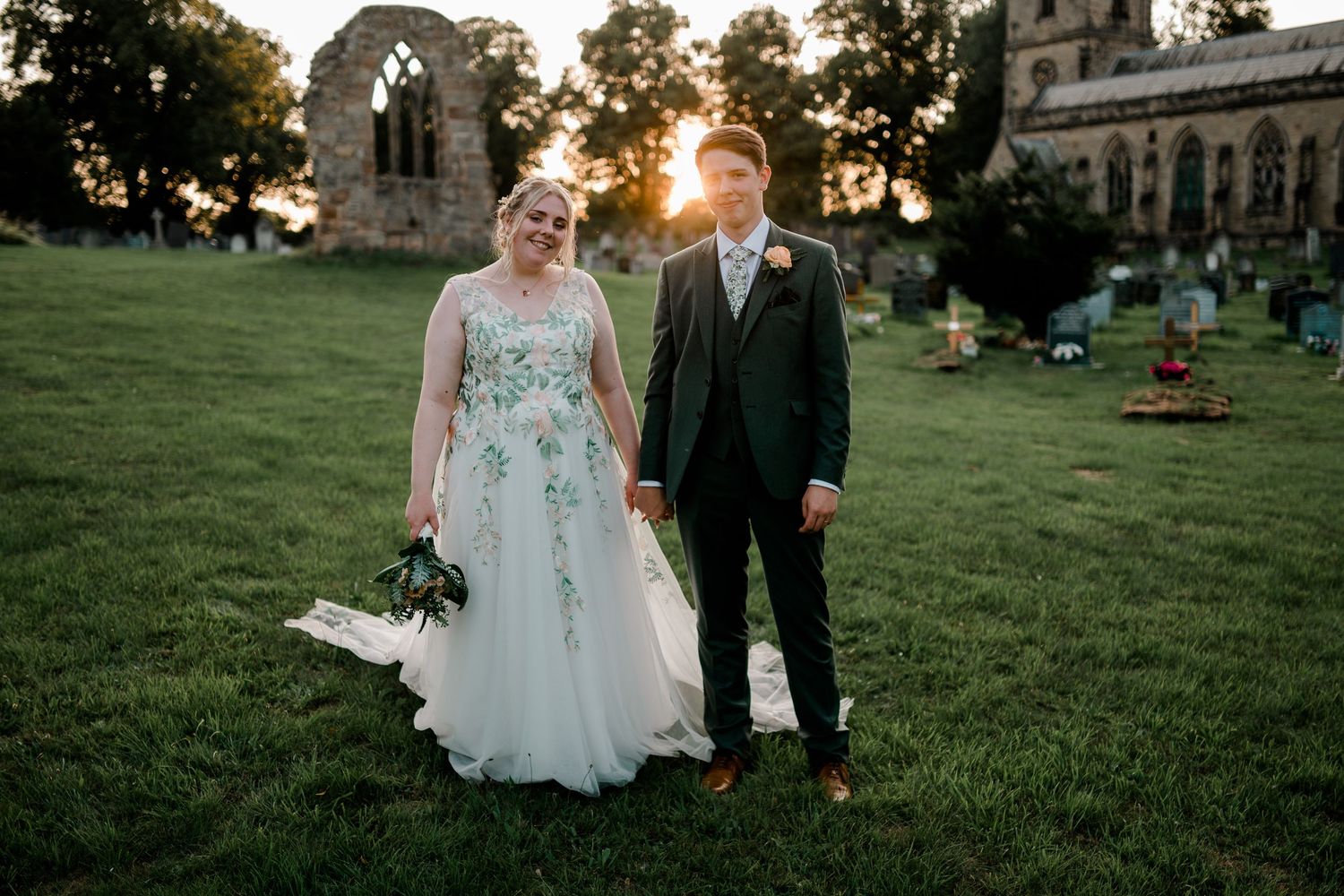 A couple walks hand in hand across a lush green lawn at sunset during their wedding celebration.