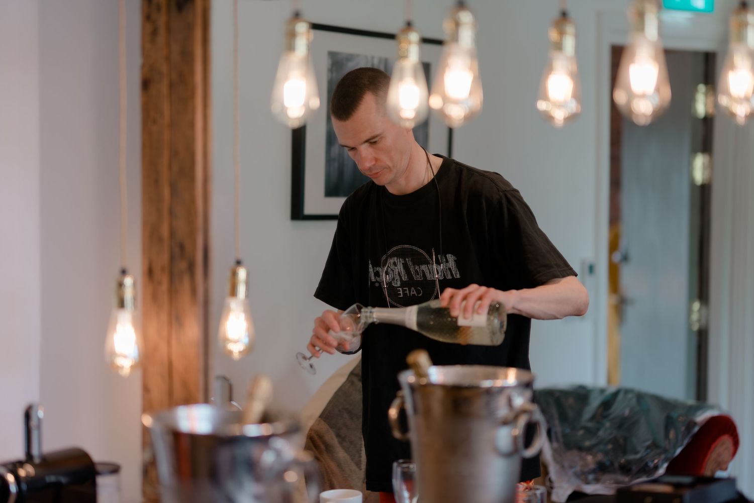 A person pours liquid into a container in a dimly lit space with hanging Edison bulbs overhead.