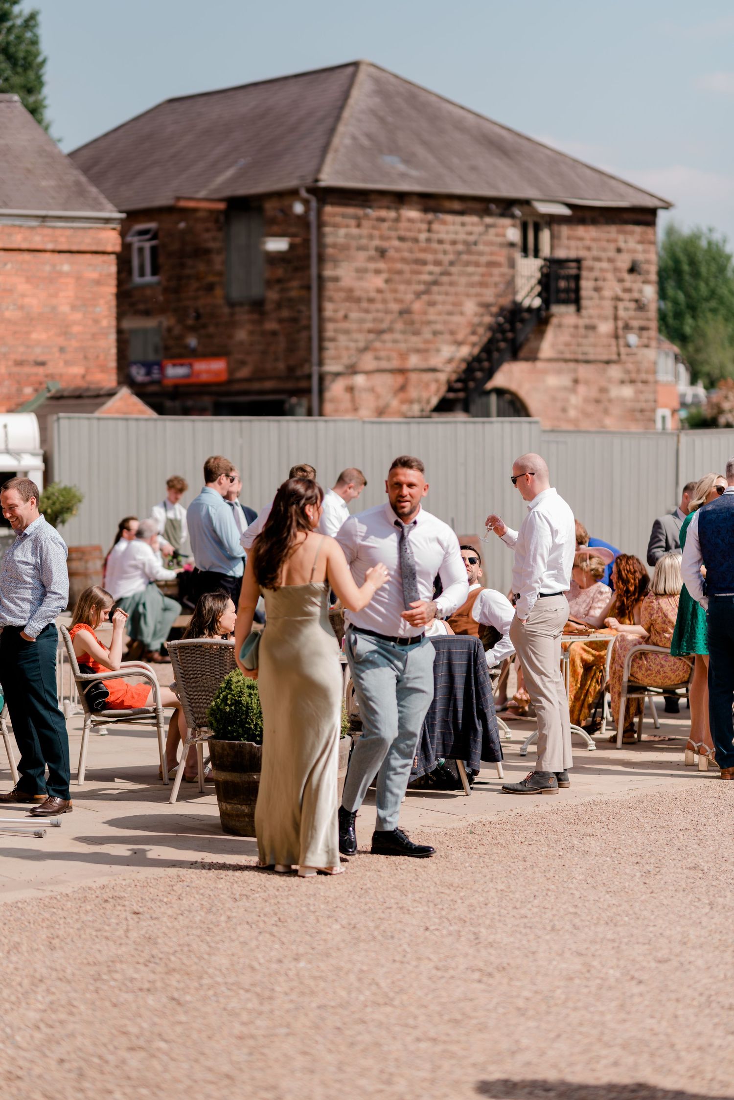 Outdoor wedding reception with guests mingling on a patio near a brick building on a sunny day.