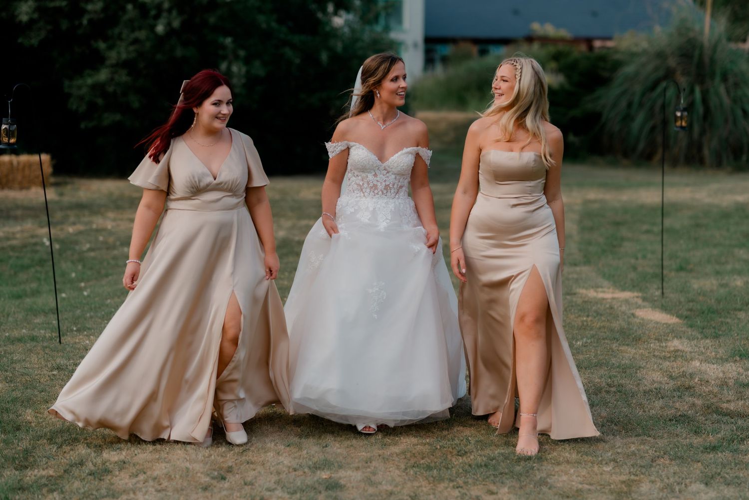 Three women in formal gowns walk together on a sunny outdoor lawn after a wedding ceremony.