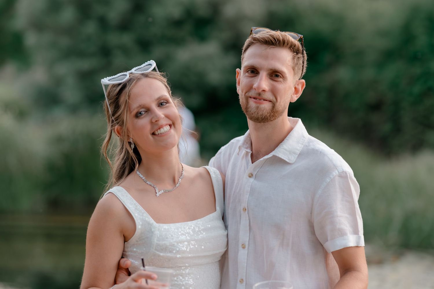 A couple in white attire poses outdoors against a natural green backdrop.