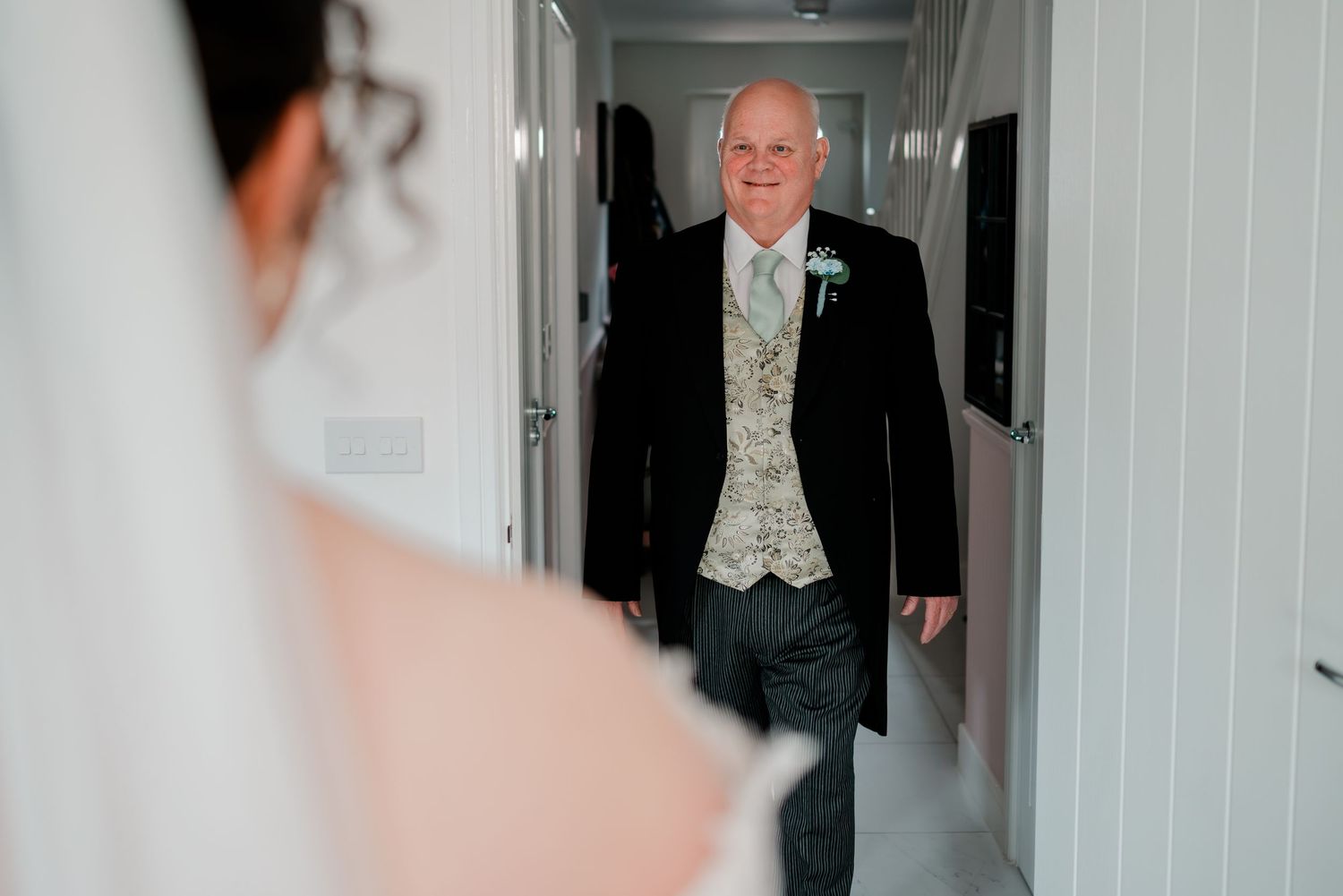 Father of the bride walking down hallway in black suit and gold waistcoat for wedding day first look.