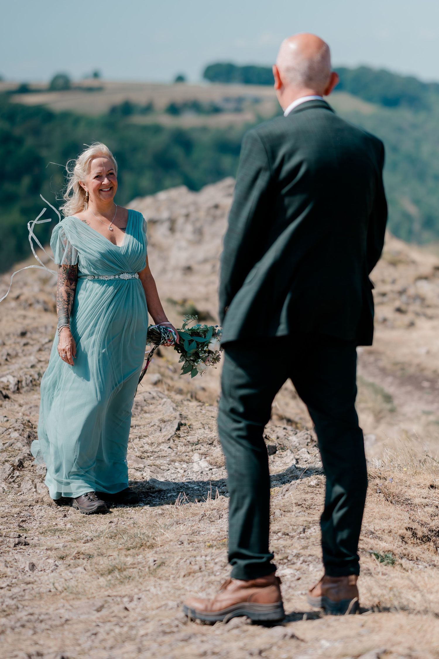 Bride in teal gown smiles at bald groom in dark suit on rocky hilltop with green hills backdrop.