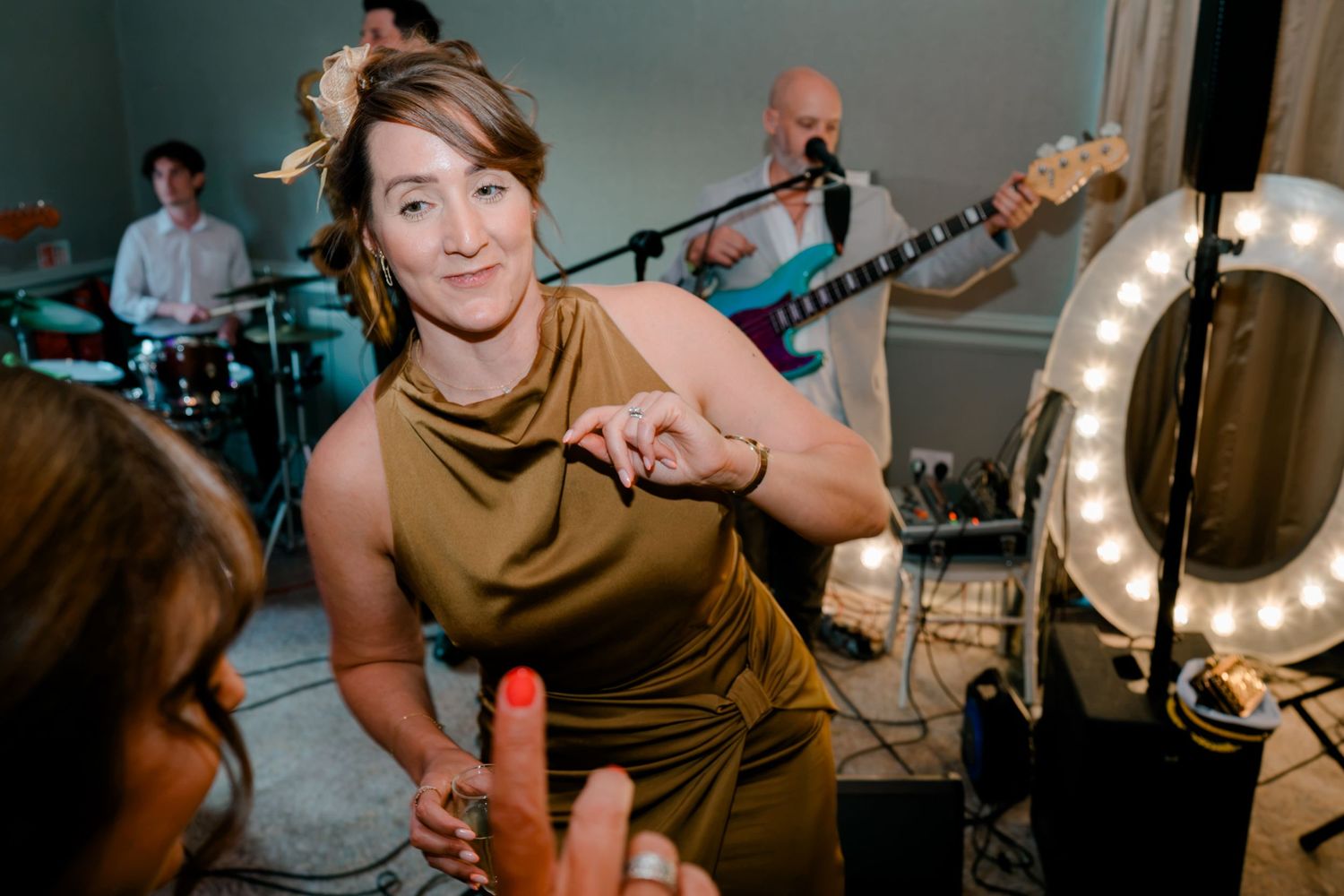 Smiling woman in a gold satin dress with a floral hair piece dances near a live band and a lighted ring mirror at a wedding reception.
