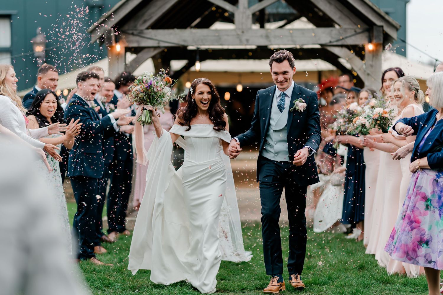 Bride and groom walk through confetti shower surrounded by cheering guests outside a barn venue.