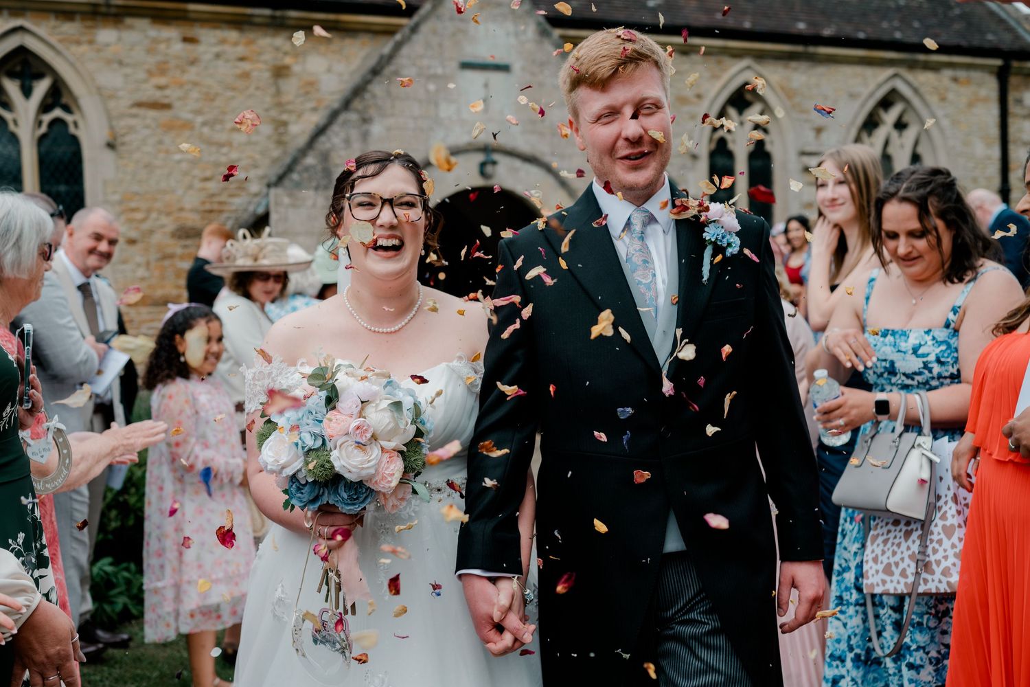 Joyful newlyweds walk through confetti shower outside stone church, bride holding pastel bouquet in white gown.