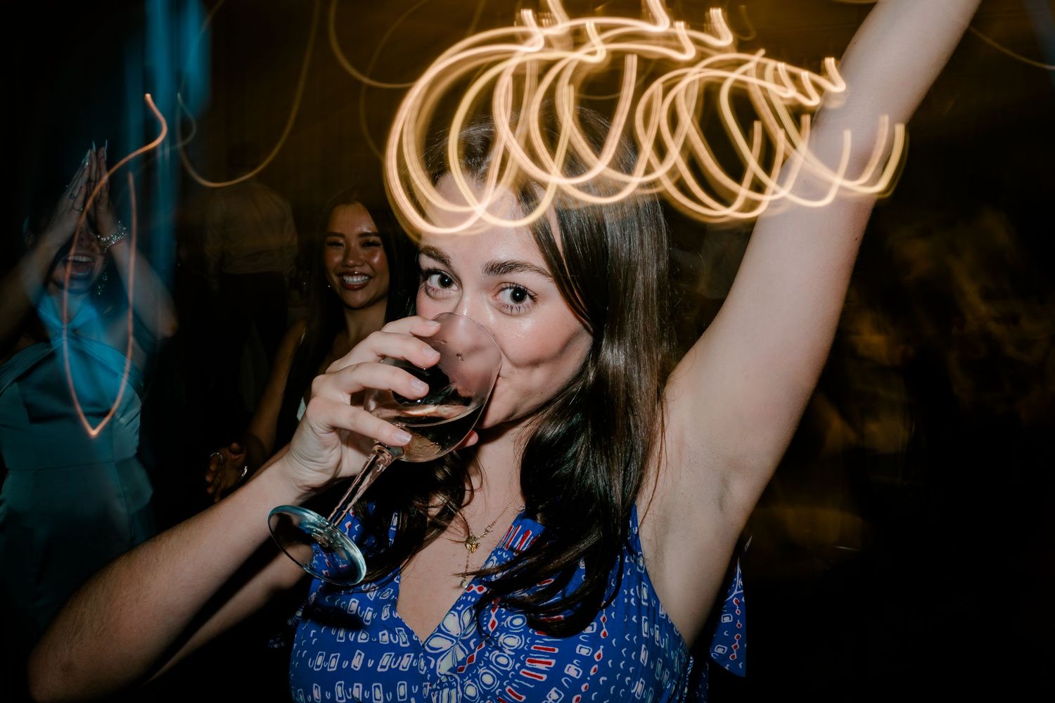 Woman dancing and drinking wine at party with glowing light trails in background.