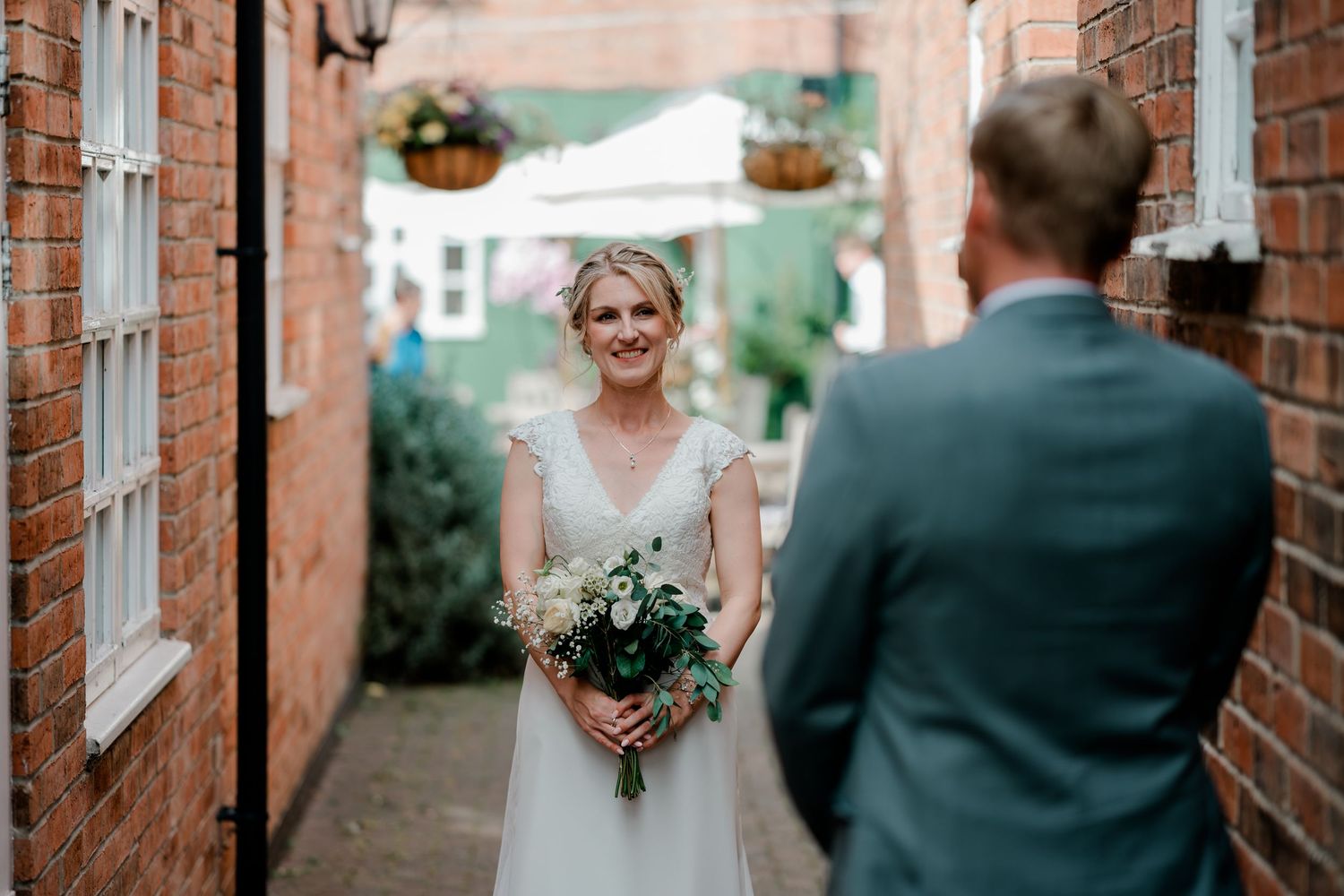 Smiling bride in white gown holding bouquet faces groom in blue suit in a brick alleyway.