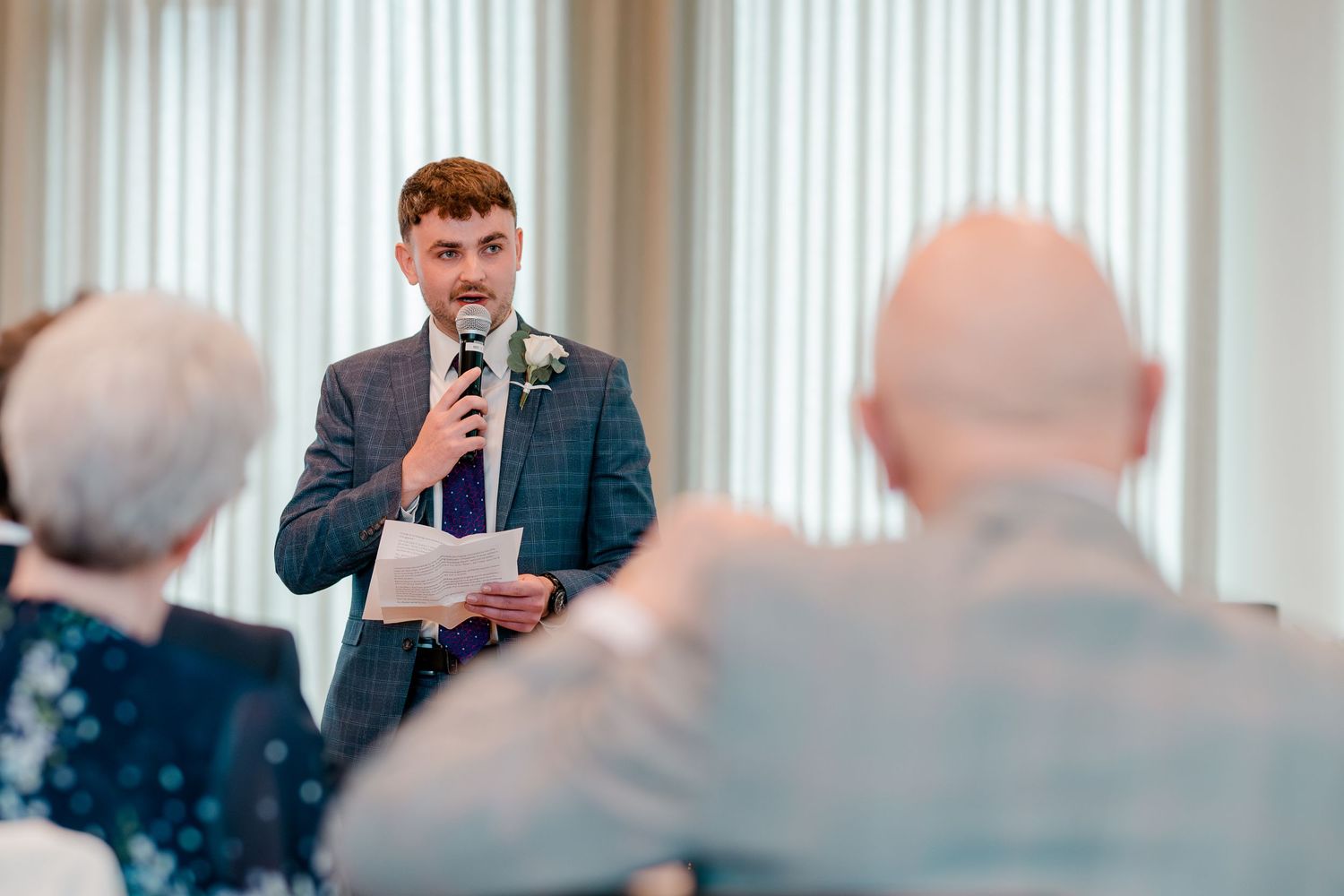 A young man in a suit delivers a speech at a wedding holding a microphone and notes.