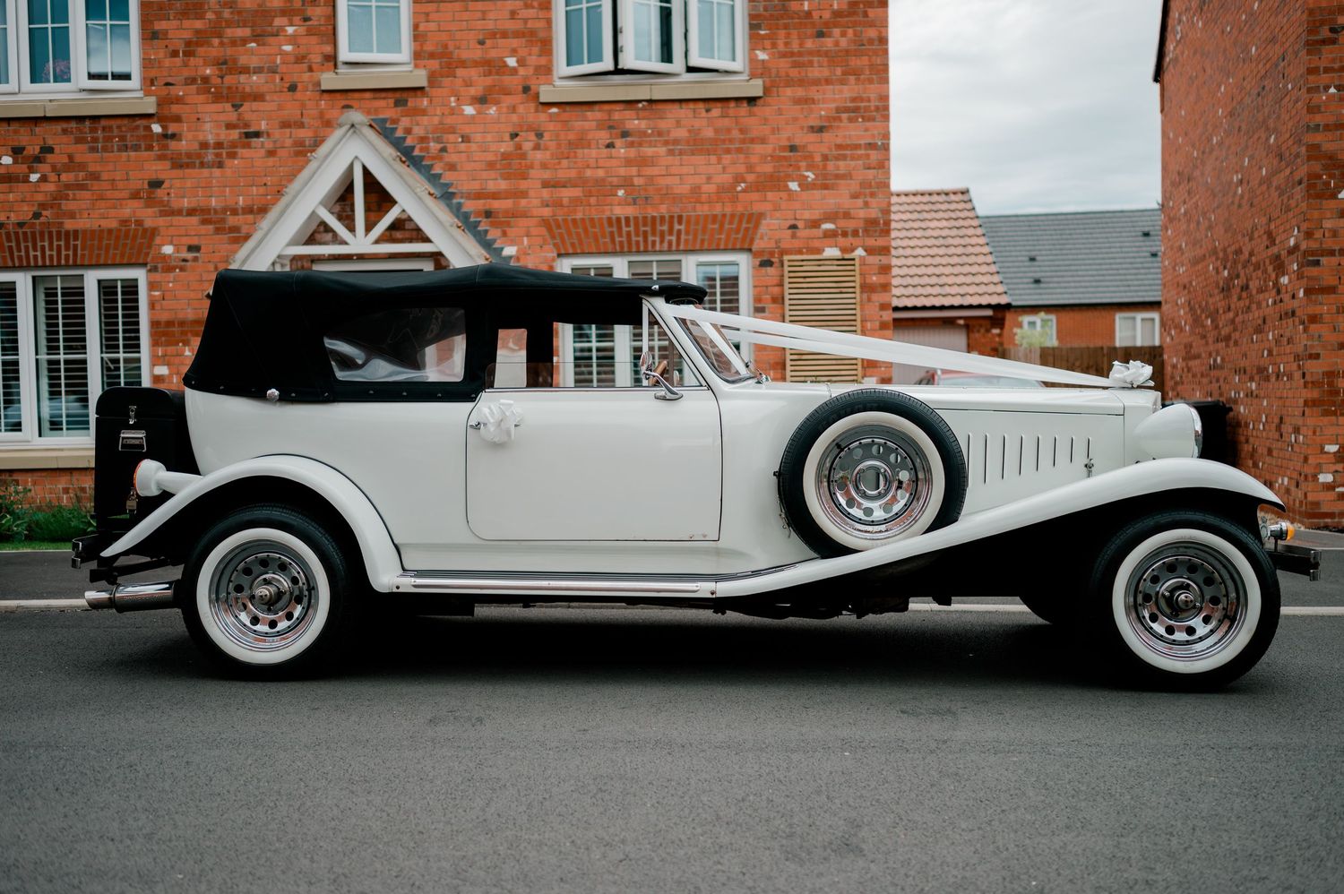 Elegant vintage white wedding car with black trim and ribbon decoration parked outside a brick building.