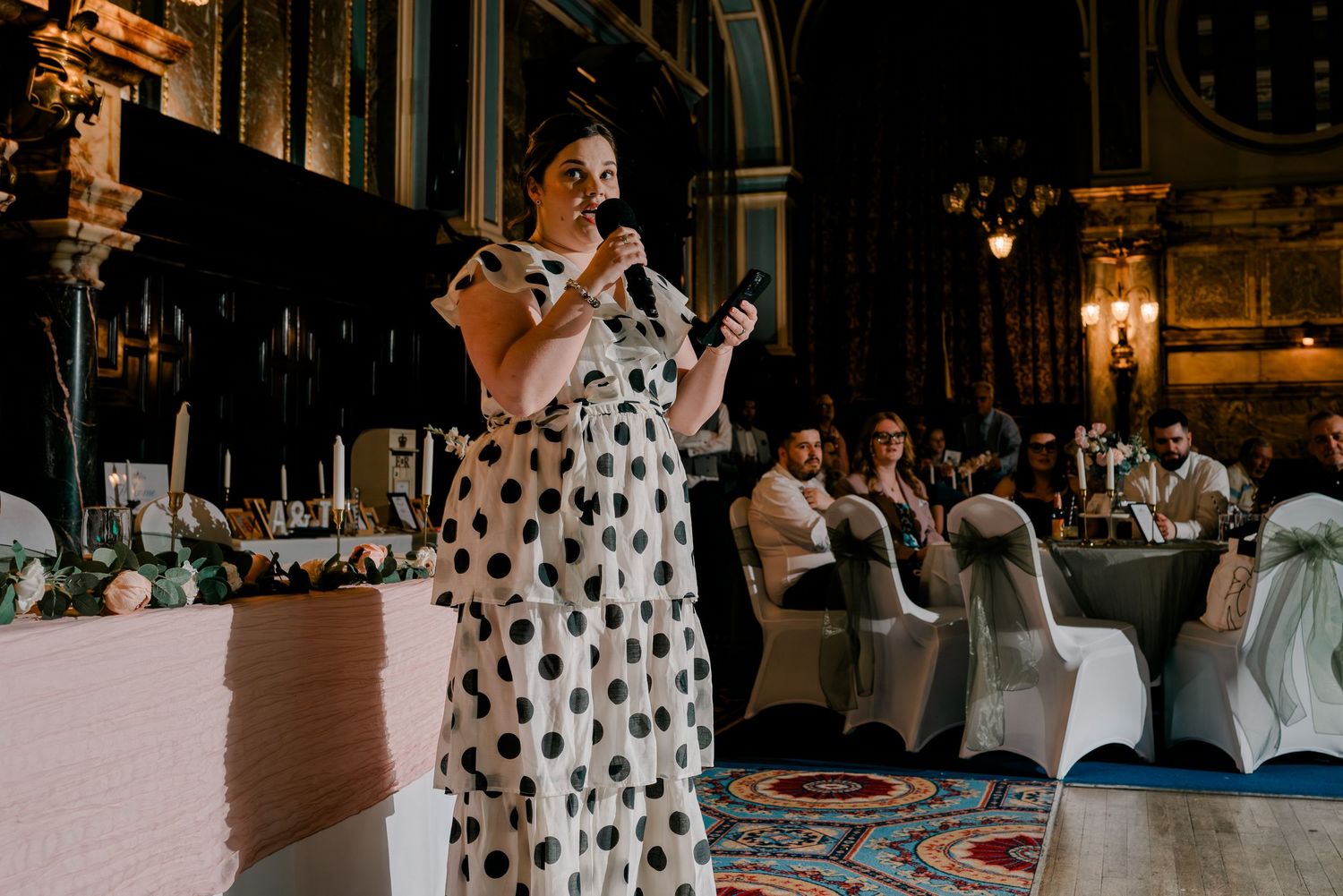 Woman in polka dot dress giving speech at wedding reception in ornate venue.