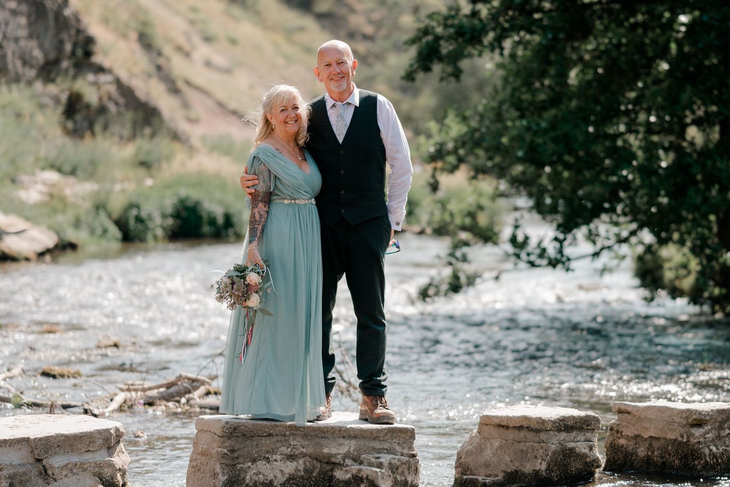 Smiling couple posing on stepping stones by a scenic river after their wedding ceremony.