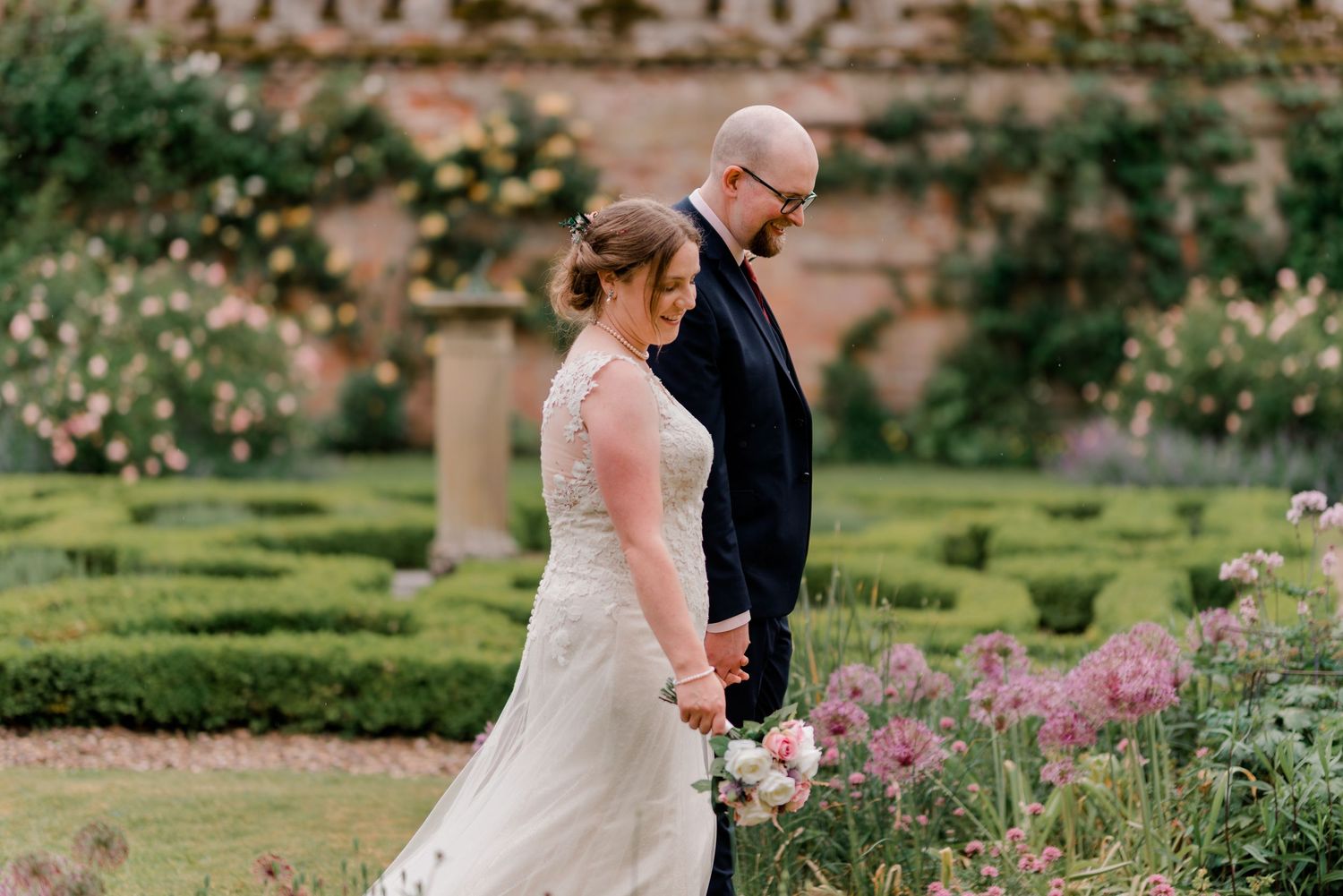 Bride and bespectacled groom walk hand in hand through beautiful rose garden of historic estate.