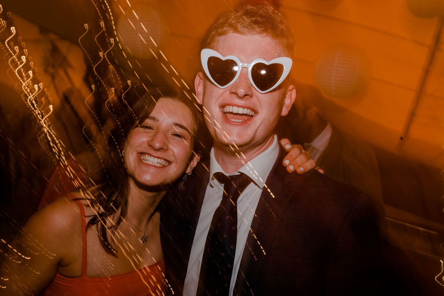 Two guests laughing at a wedding reception, one wearing novelty white heart-shaped sunglasses.