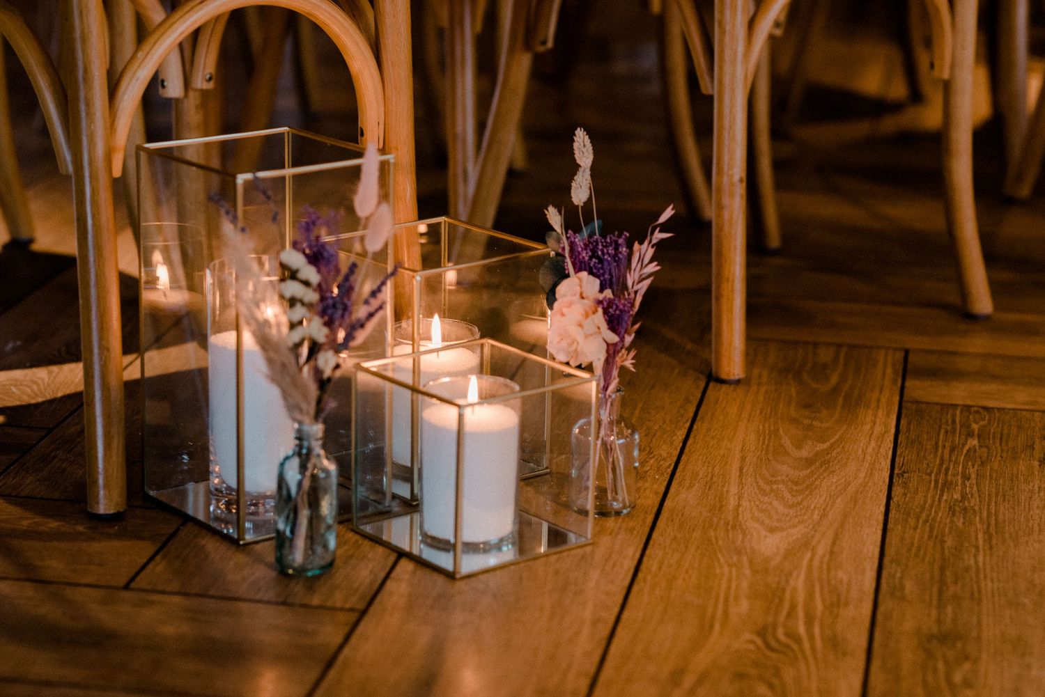 Elegant wedding aisle decor with pillar candles in glass holders and dried floral arrangements on herringbone floor.