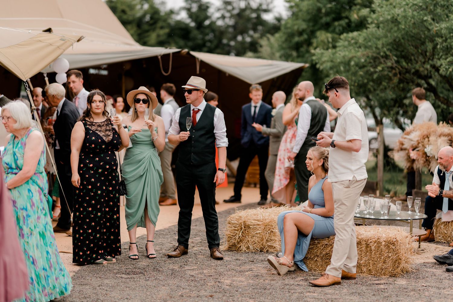 Guests gather at an outdoor rustic wedding reception with hay bales and tent canopies in a garden setting.