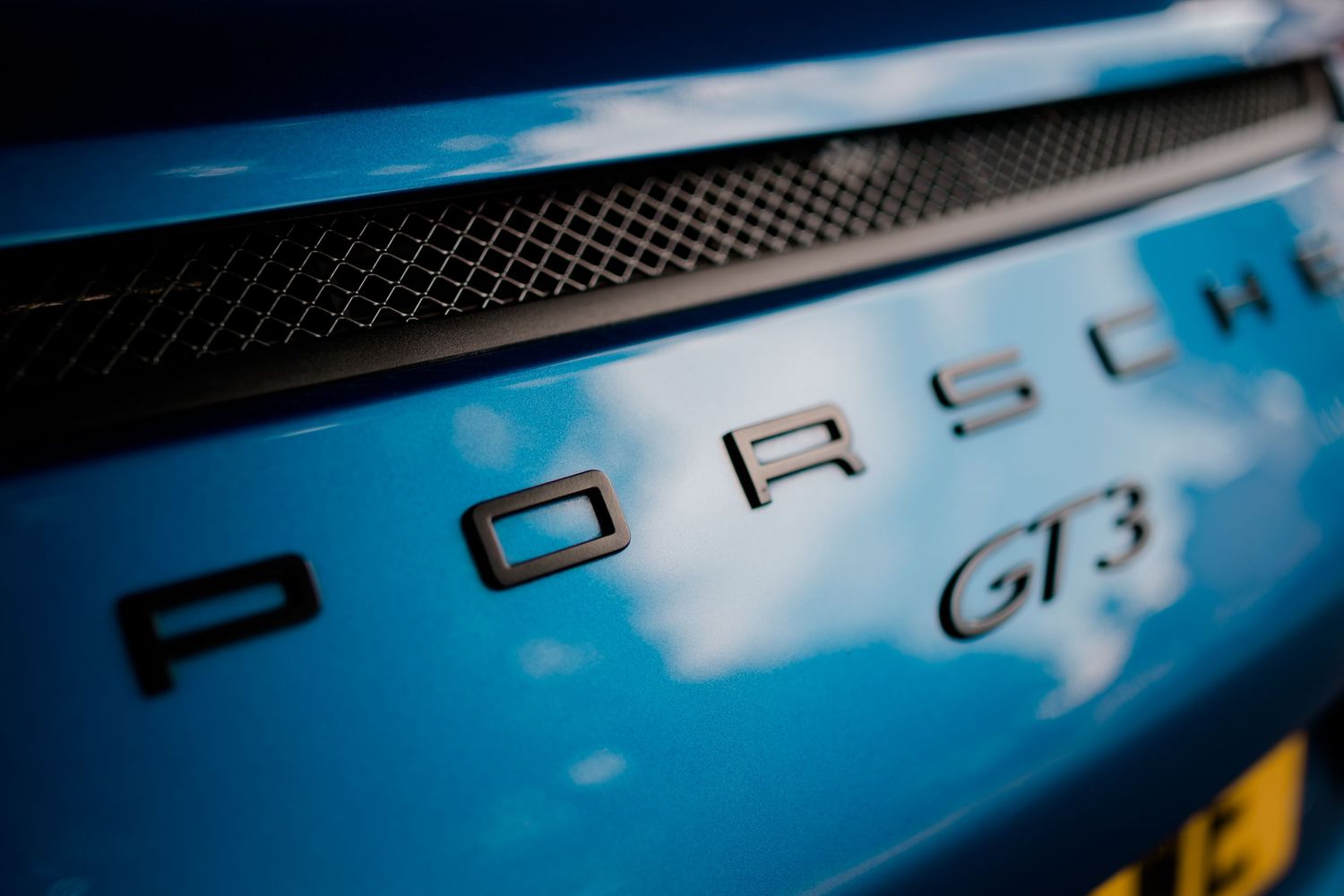 Close-up view of a black Porsche badge and lettering against a metallic car surface under blue sky.