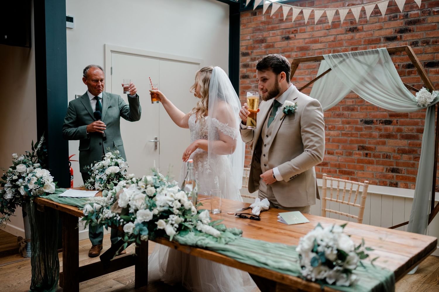 Bride and groom toasting champagne at wedding signing table with white floral arrangements and bunting decor.