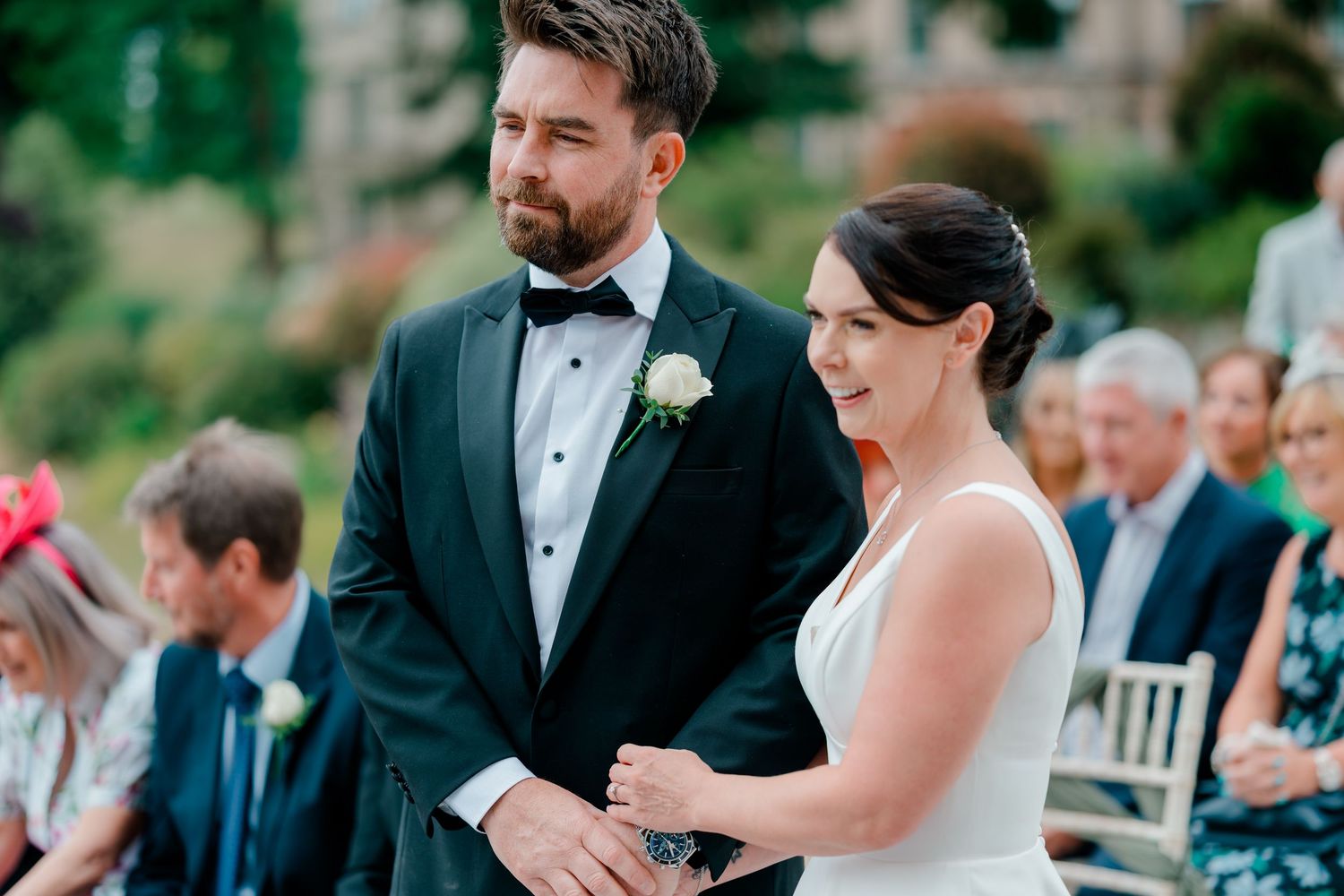 Groom in a black tuxedo with bow tie and white boutonniere holds hands with a bride in a white dress during an outdoor wedding ceremony.