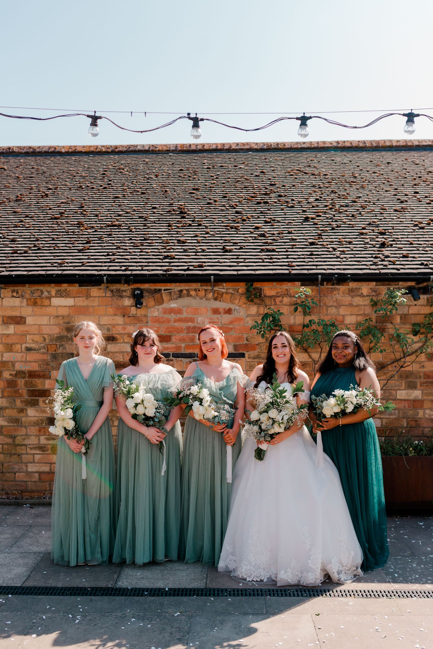 Bridal party in sage green dresses posing against brick wall with white floral bouquets.