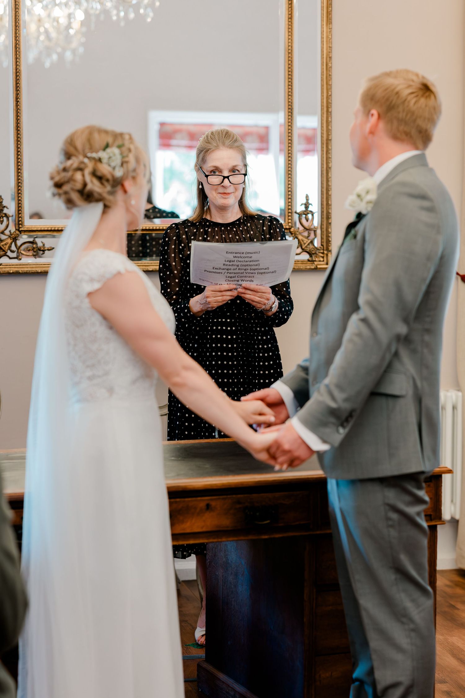Bride and groom holding hands during wedding ceremony while officiant reads vows in elegant venue.