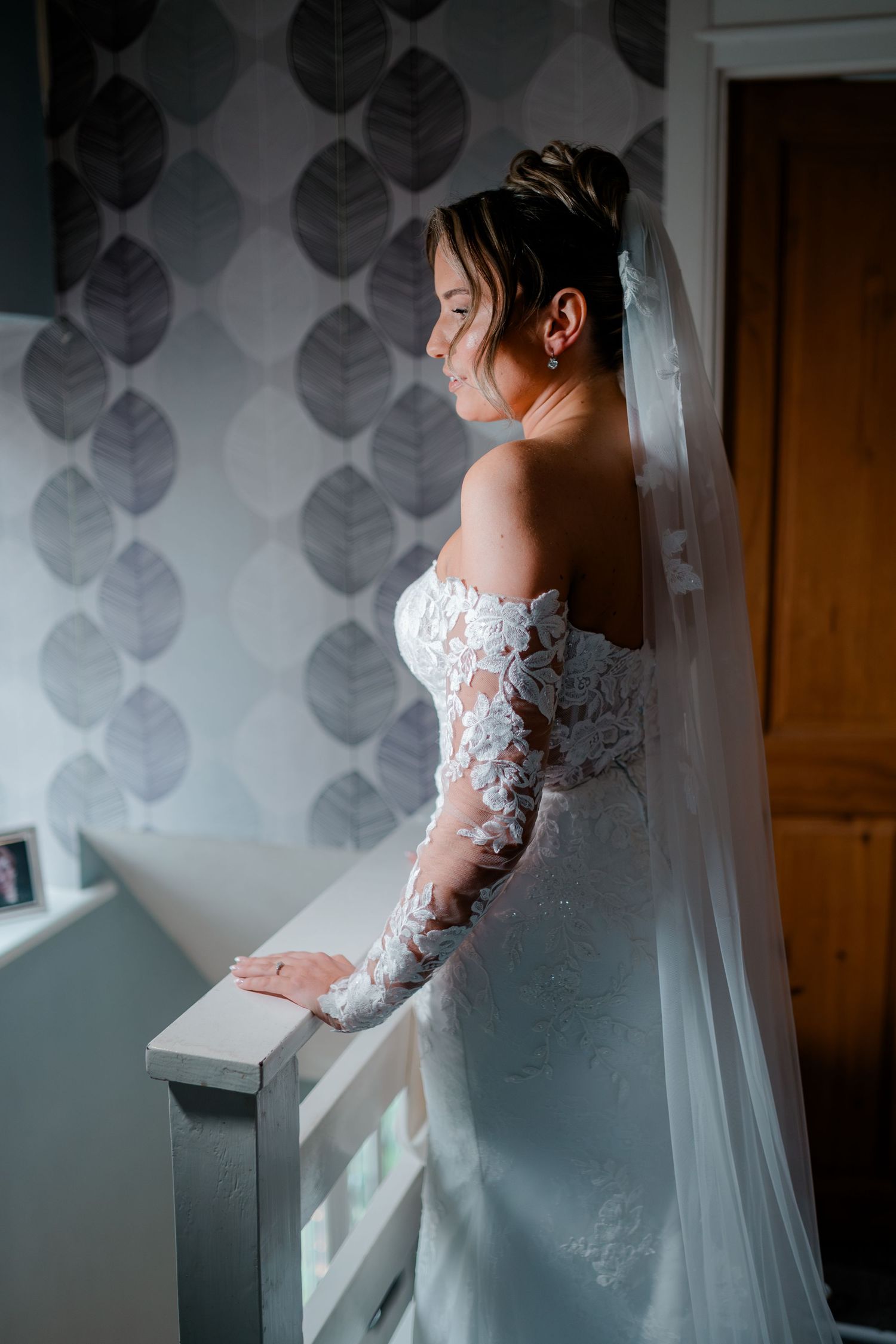Bride in lace off-shoulder wedding dress and veil stands by staircase railing, bathed in natural light.