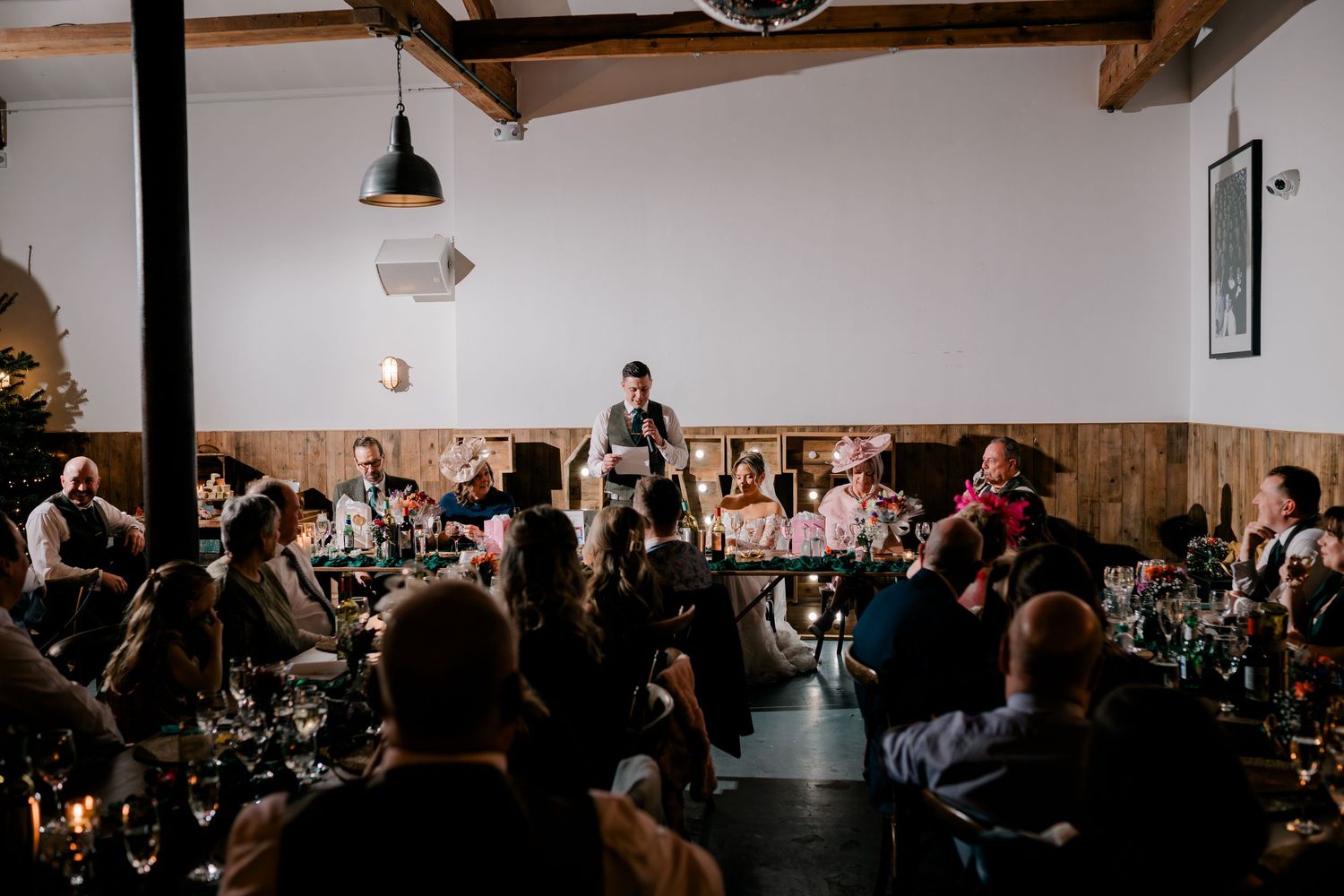 Wedding guests seated at decorated tables listen attentively to a speech at a rustic venue.