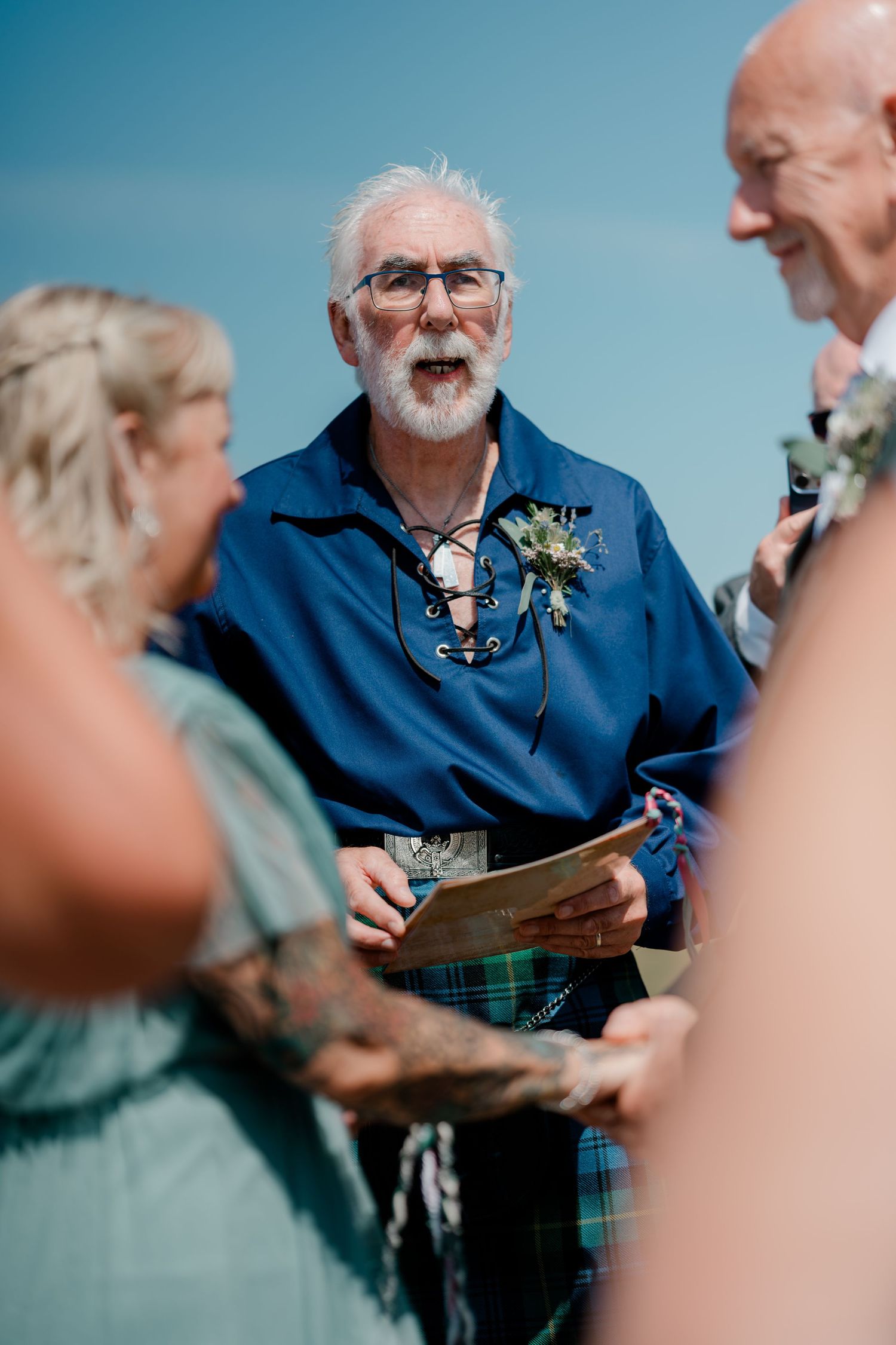 Elderly officiant in blue Scottish kilt outfit reads vows at outdoor wedding ceremony under clear blue sky.