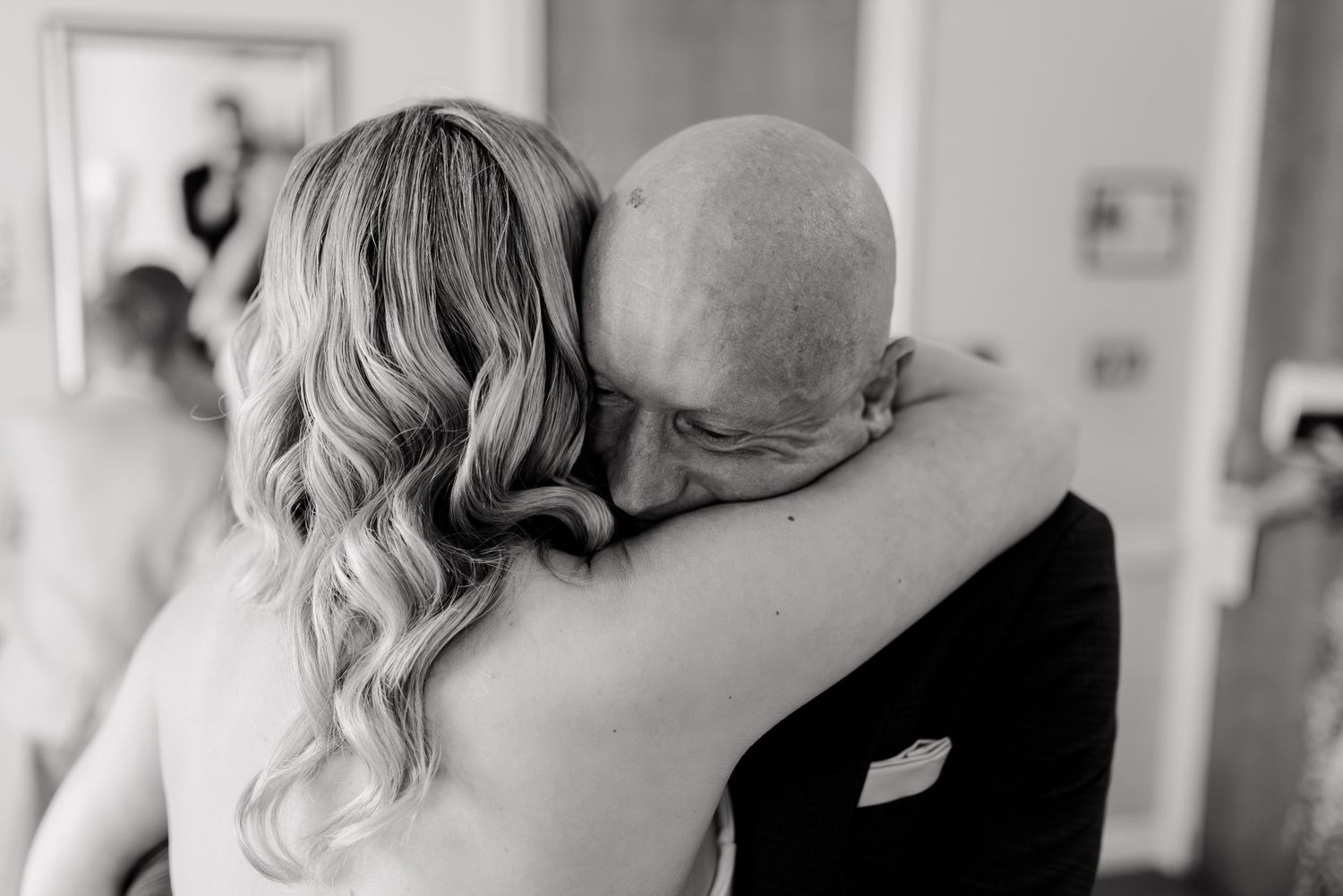Emotional black and white photo of a bride embracing a bald man during wedding preparations.