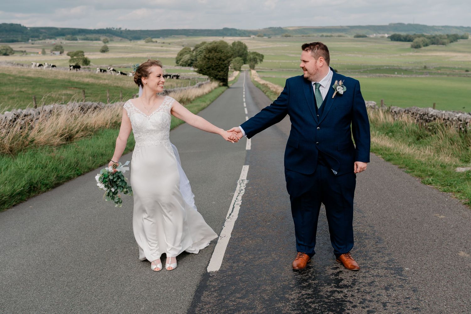 Bride and groom hold hands on a quiet rural road surrounded by rolling green countryside fields.