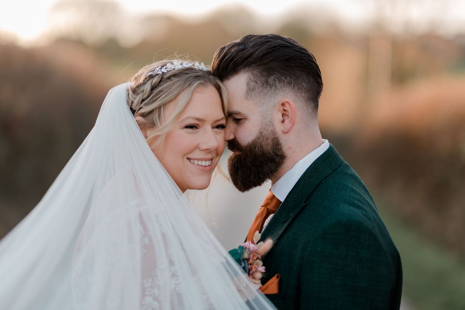 A tender moment between newlyweds with a white veil and emerald green suit in golden sunset light.