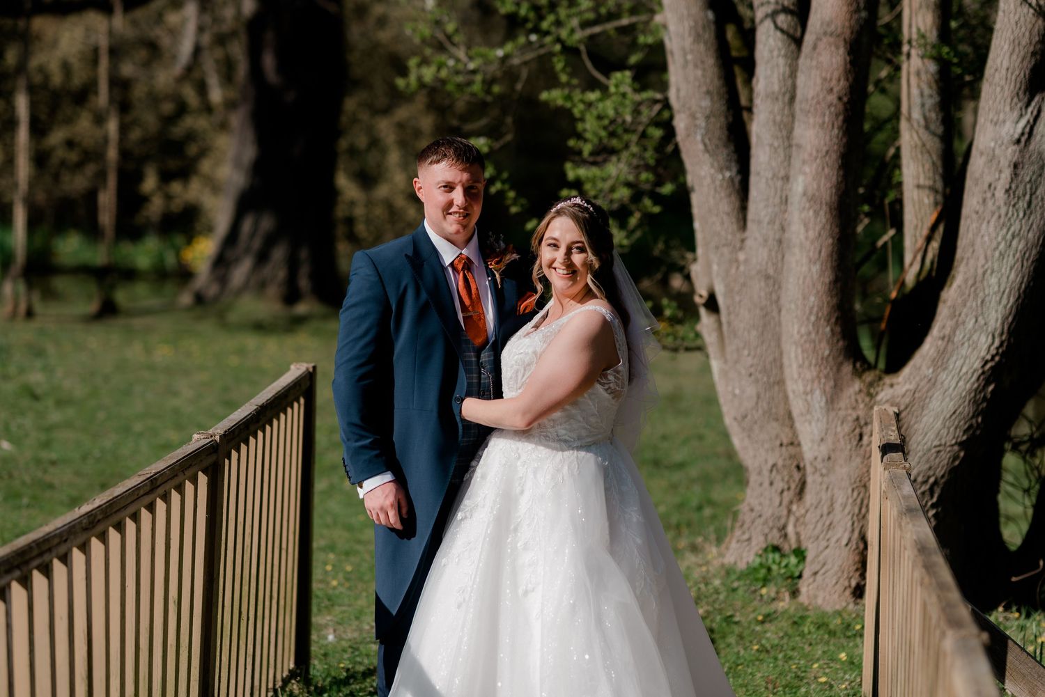 Couple walks along rustic wooden bridge surrounded by tall trees during outdoor wedding photoshoot.