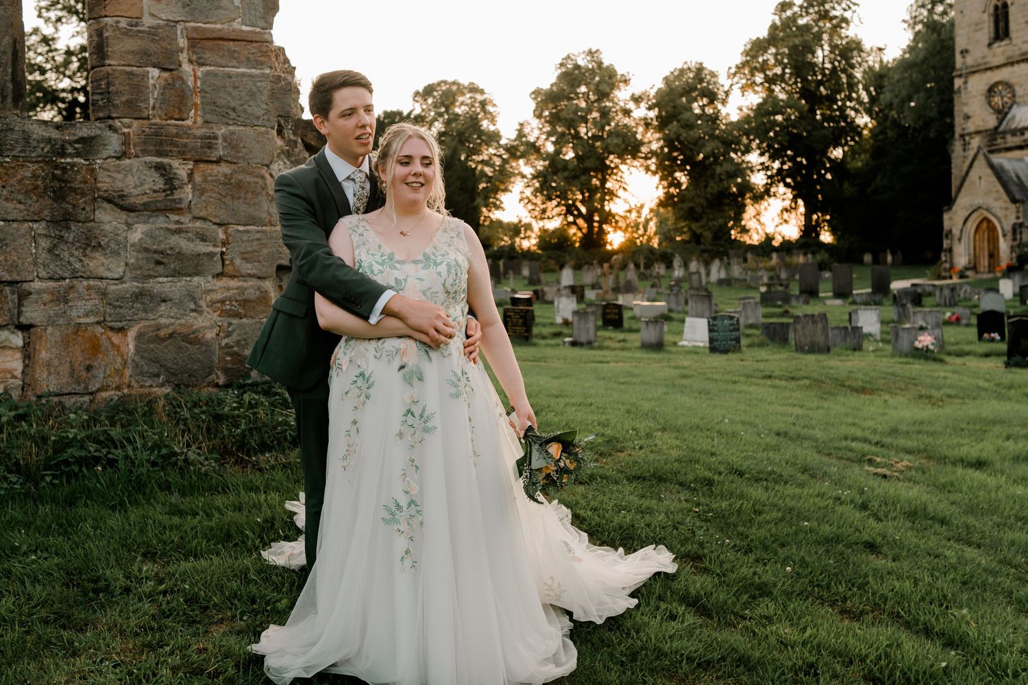 Bride in white dress poses in front of stone church during golden hour sunset.
