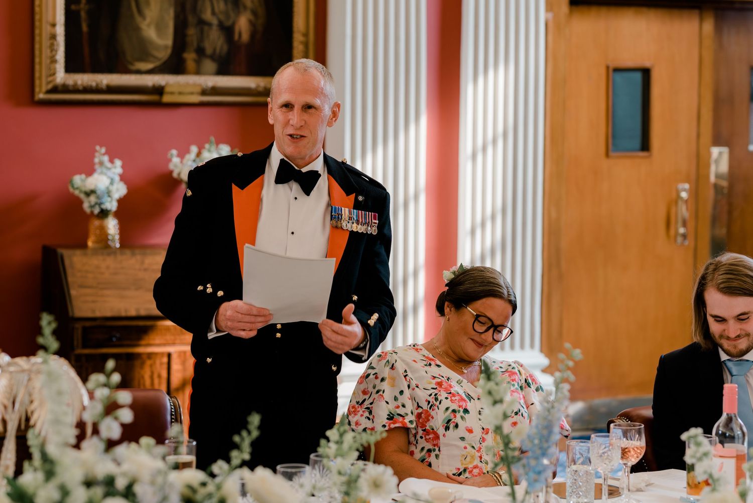 Wedding guests sit at elegantly decorated tables during a formal dinner reception in an orange-walled dining room.