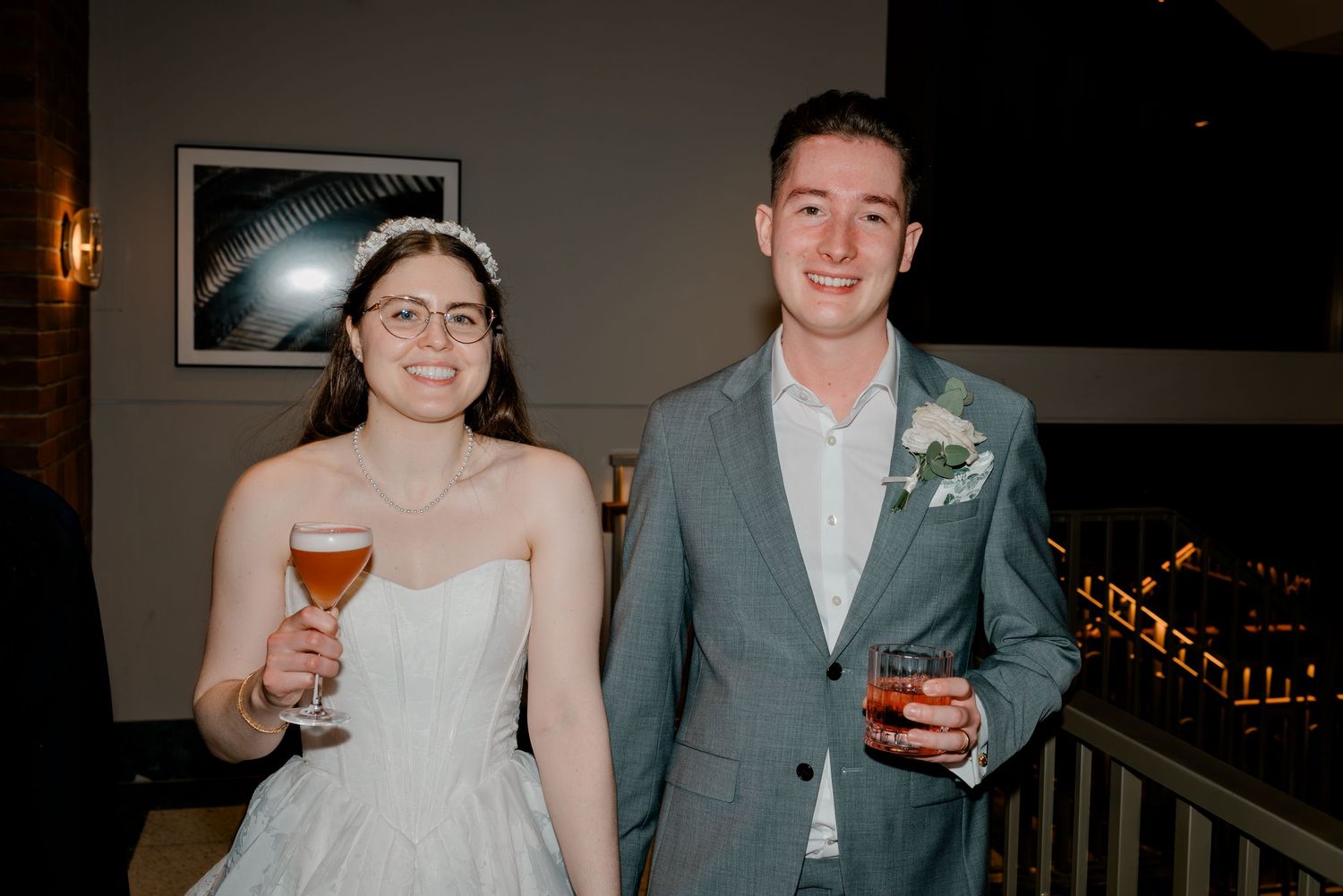 Bride in white dress and groom in gray suit smiling and holding drinks at wedding reception.