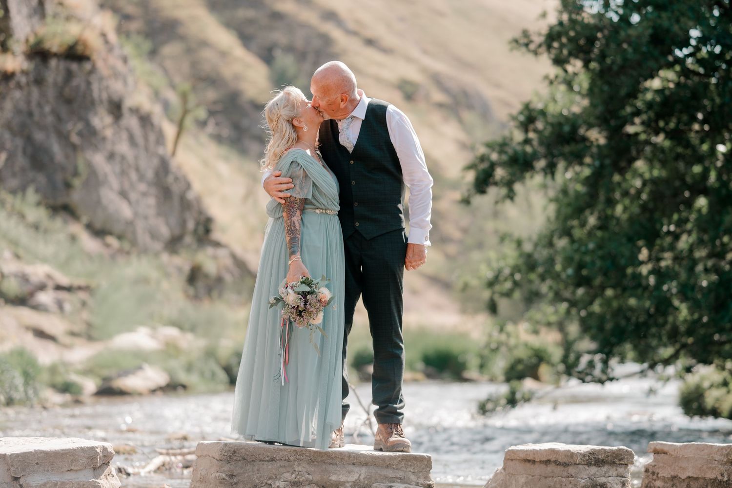Couple sharing a kiss outdoors near a river, woman in mint green gown holding bouquet, man in dark vest.