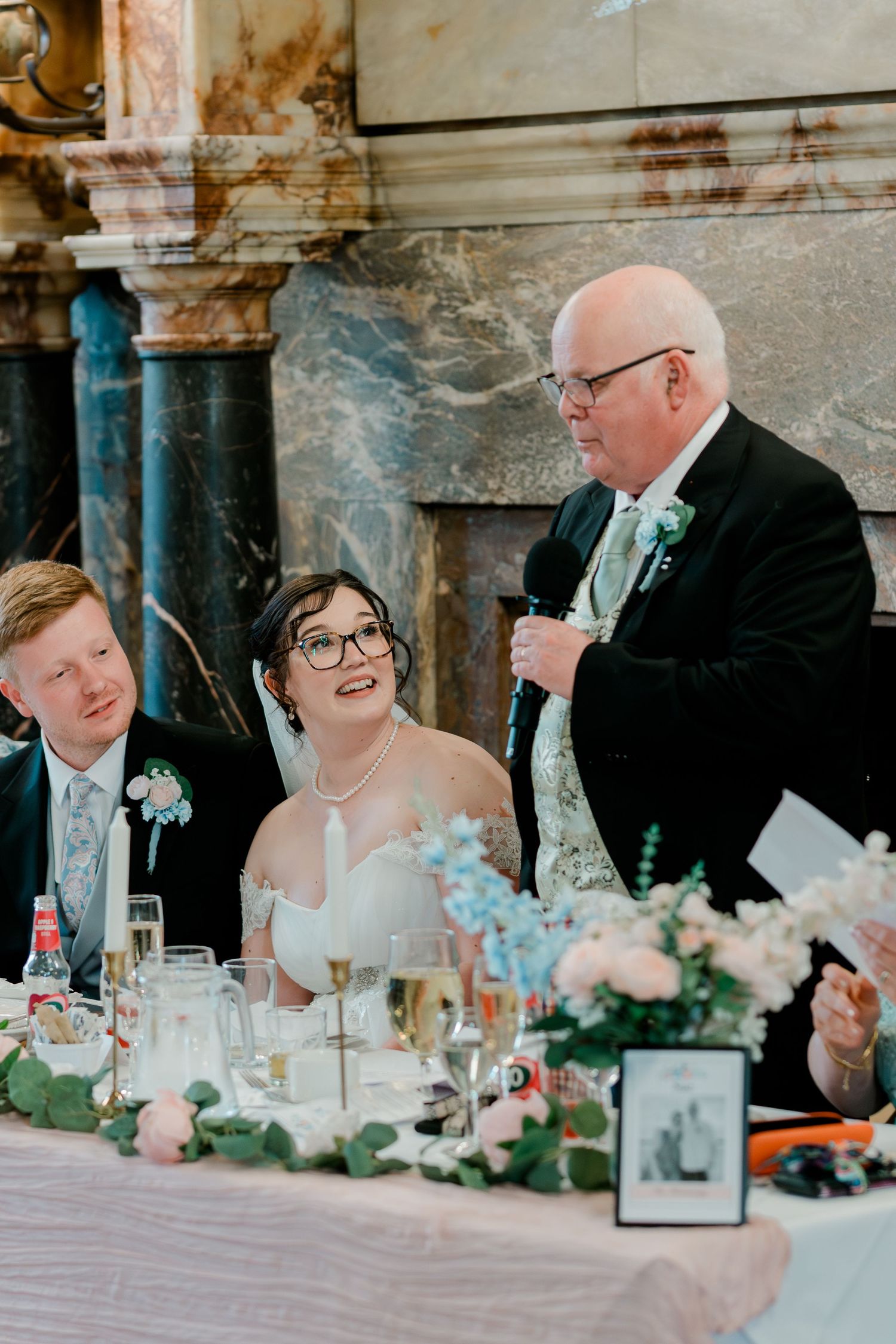 Father of bride giving speech at wedding reception as couple listens at decorated head table.