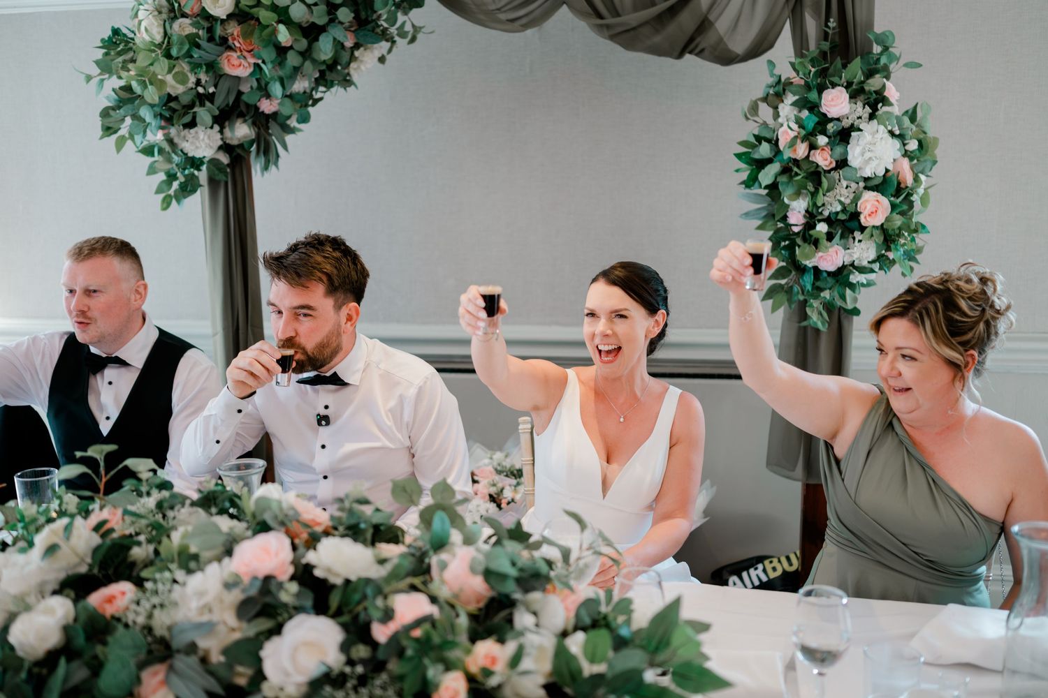 Wedding guests raise glasses in a toast at a reception table decorated with pink and white floral centerpieces and greenery garlands.