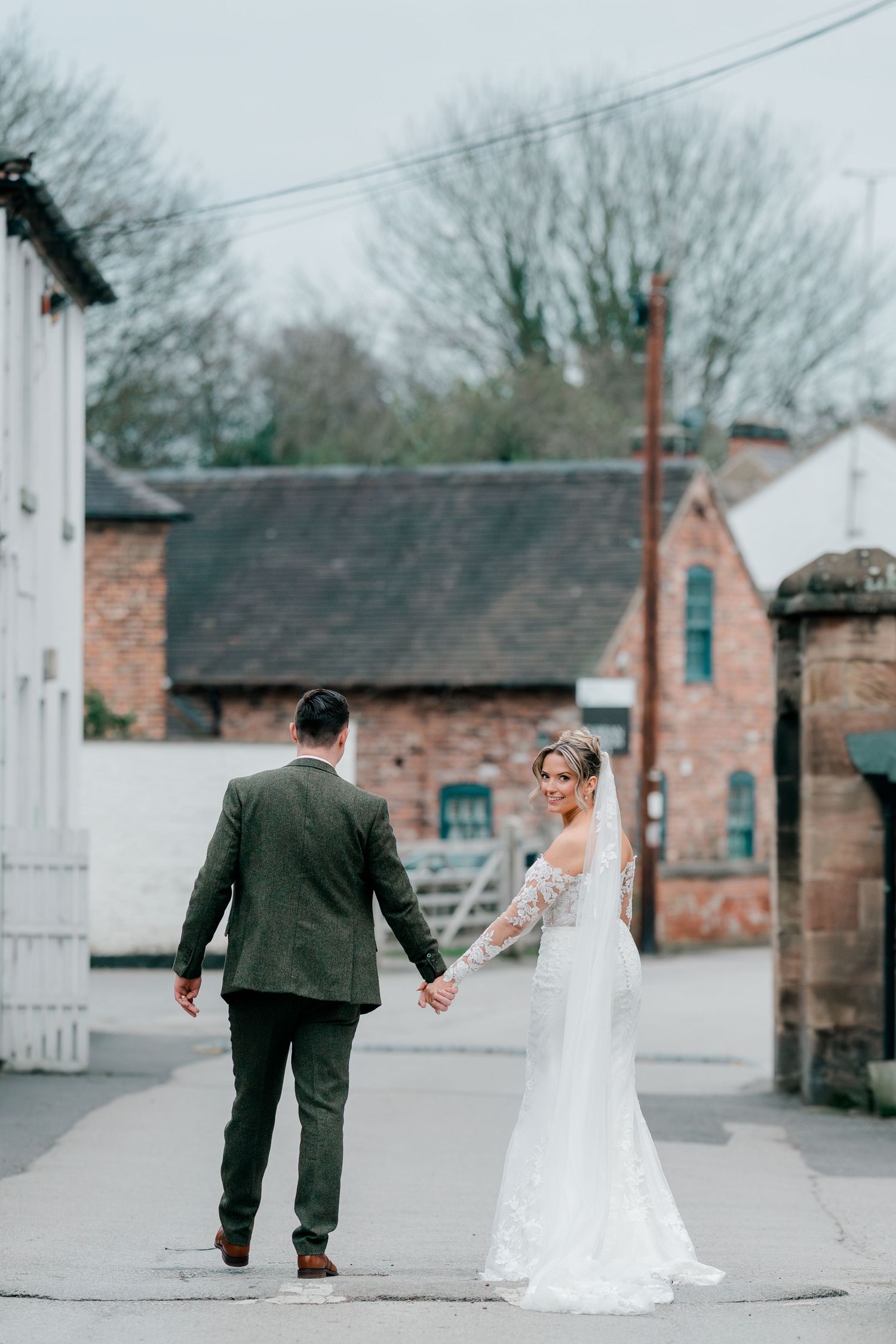 Bride in lace gown and veil holds grooms hand while looking back on a quiet street.