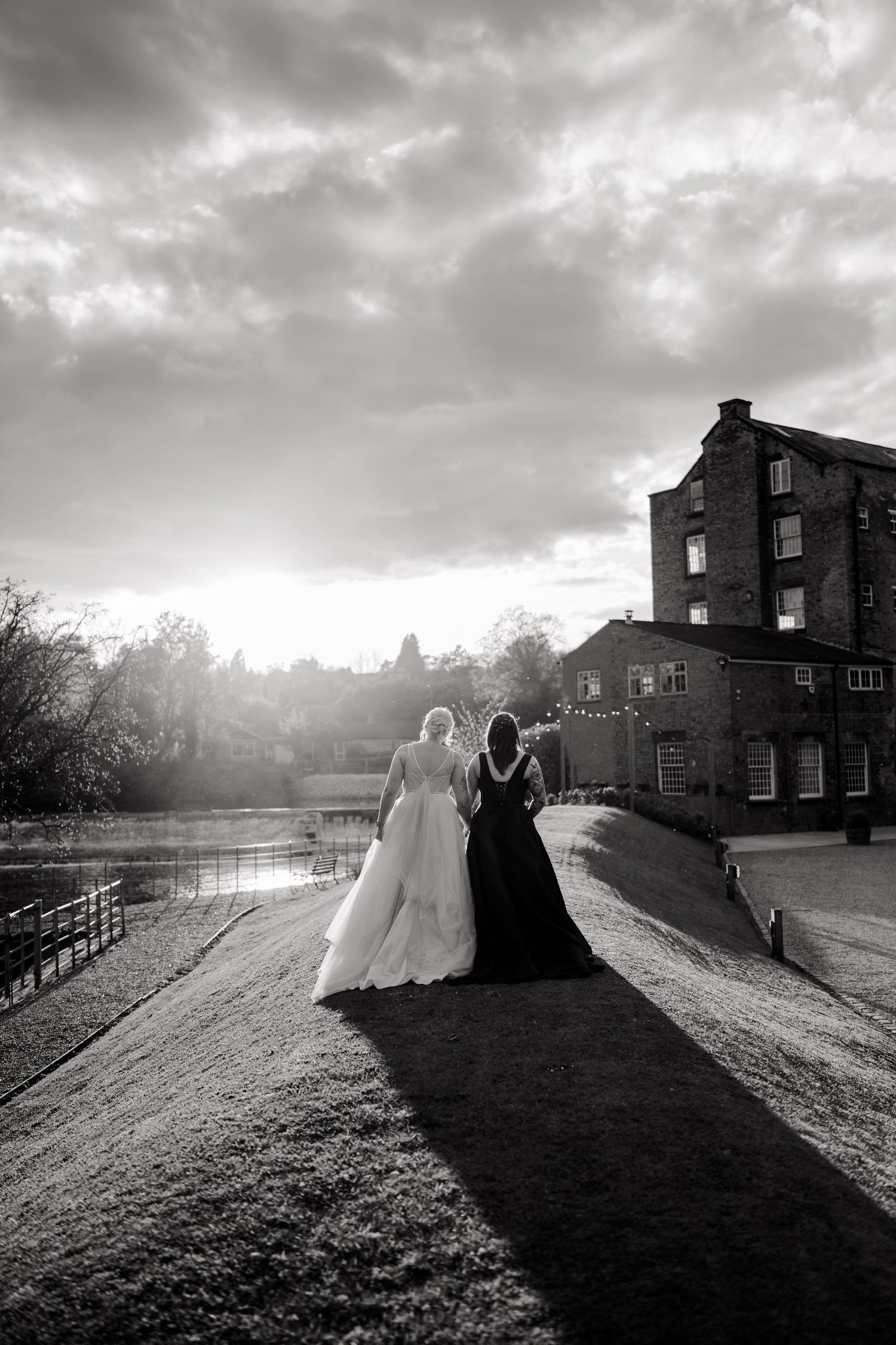 Black and white photo of two brides walking toward sunset beside historic mill building and lake.