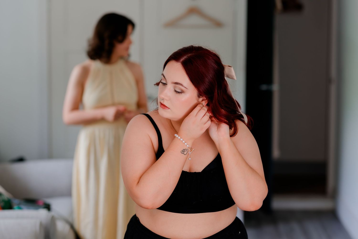 Someone in a black top adjusts their jewelry while getting ready, with another person in a yellow dress visible in background.