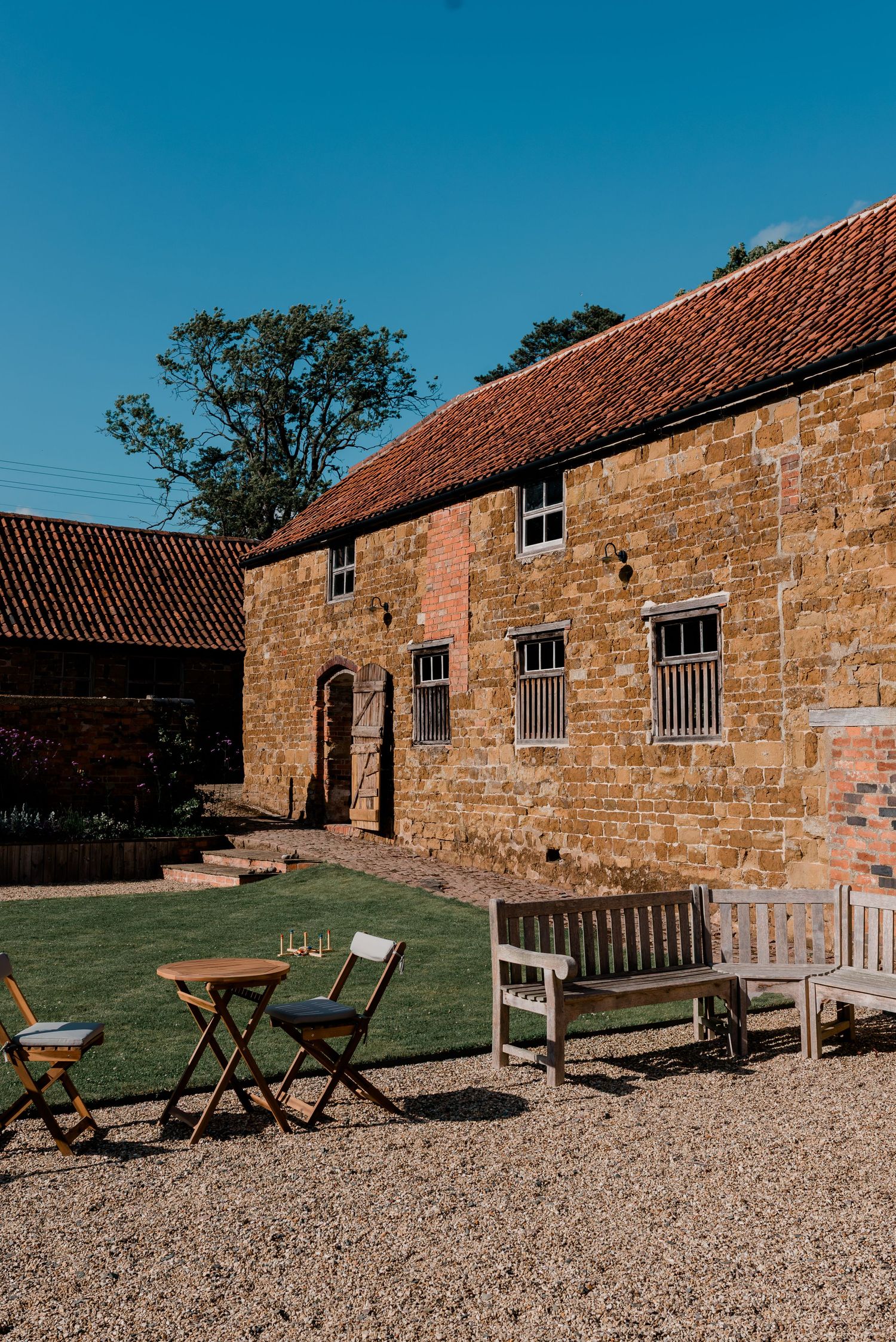 Rustic stone barns with red tile roofs and wooden outdoor furniture set on gravel courtyard on sunny day.