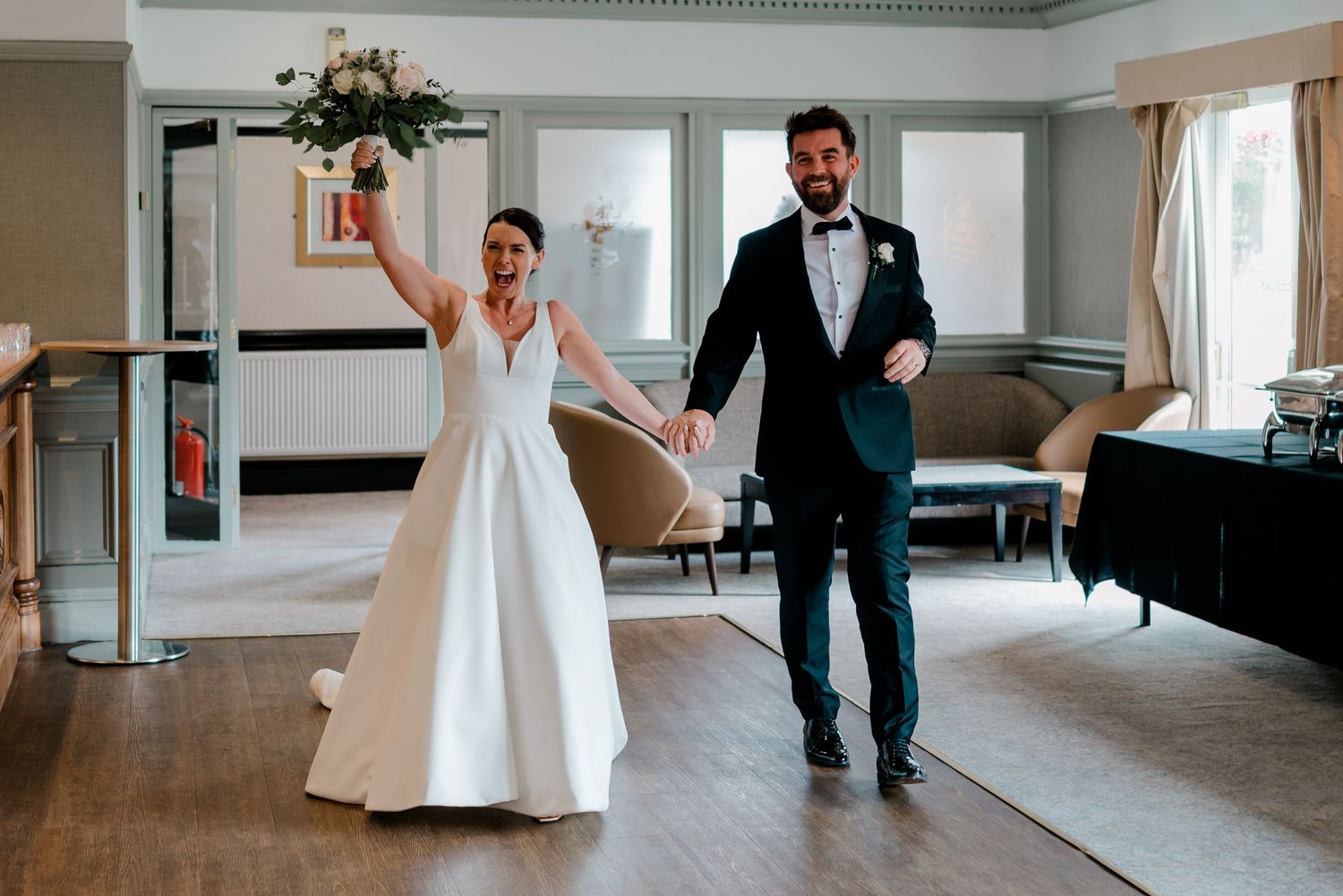 Joyful bride in a white gown raising her bouquet while holding hands with a groom in a navy suit as they enter their wedding reception.