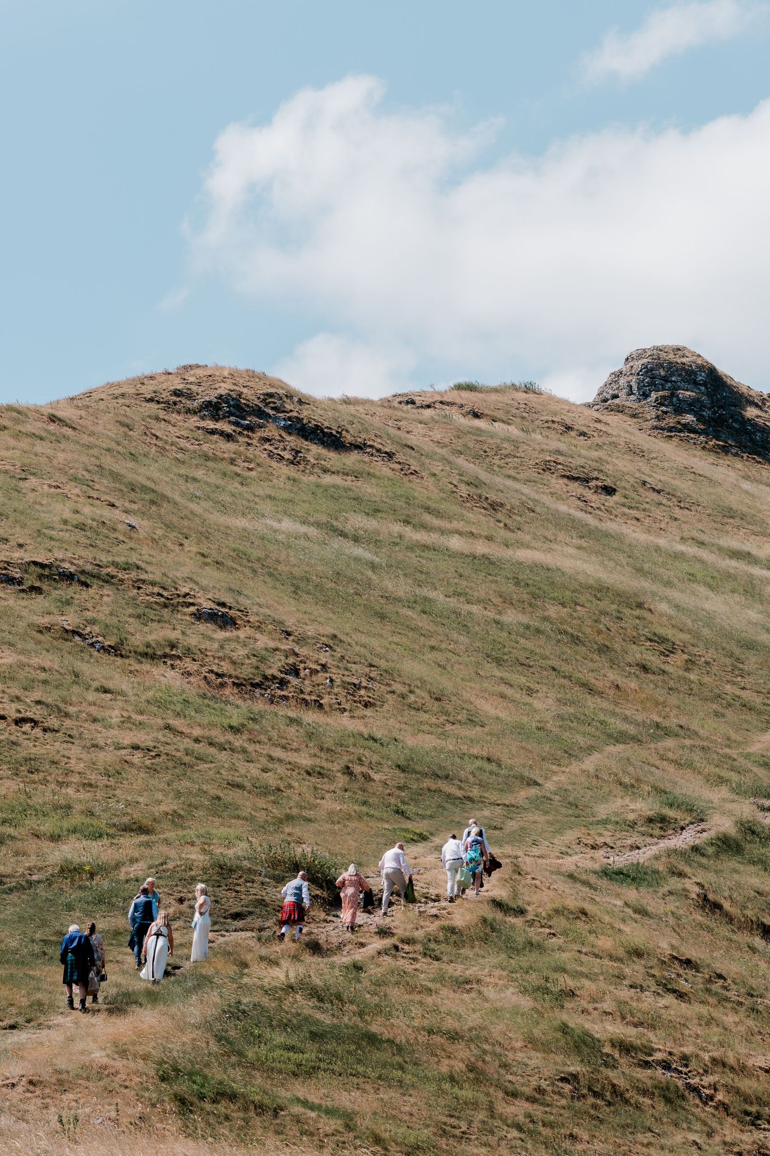 Wedding guests hike up a grassy mountain trail toward the rocky summit.